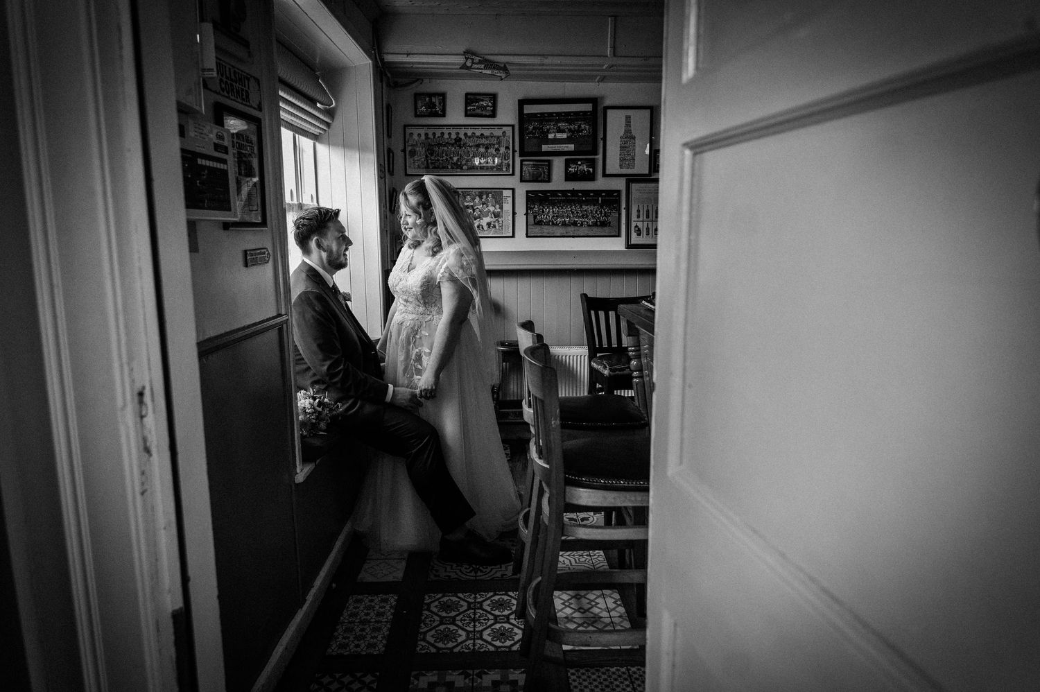 Black and white wedding photo of bride and groom sharing intimate moment by window in rustic venue.