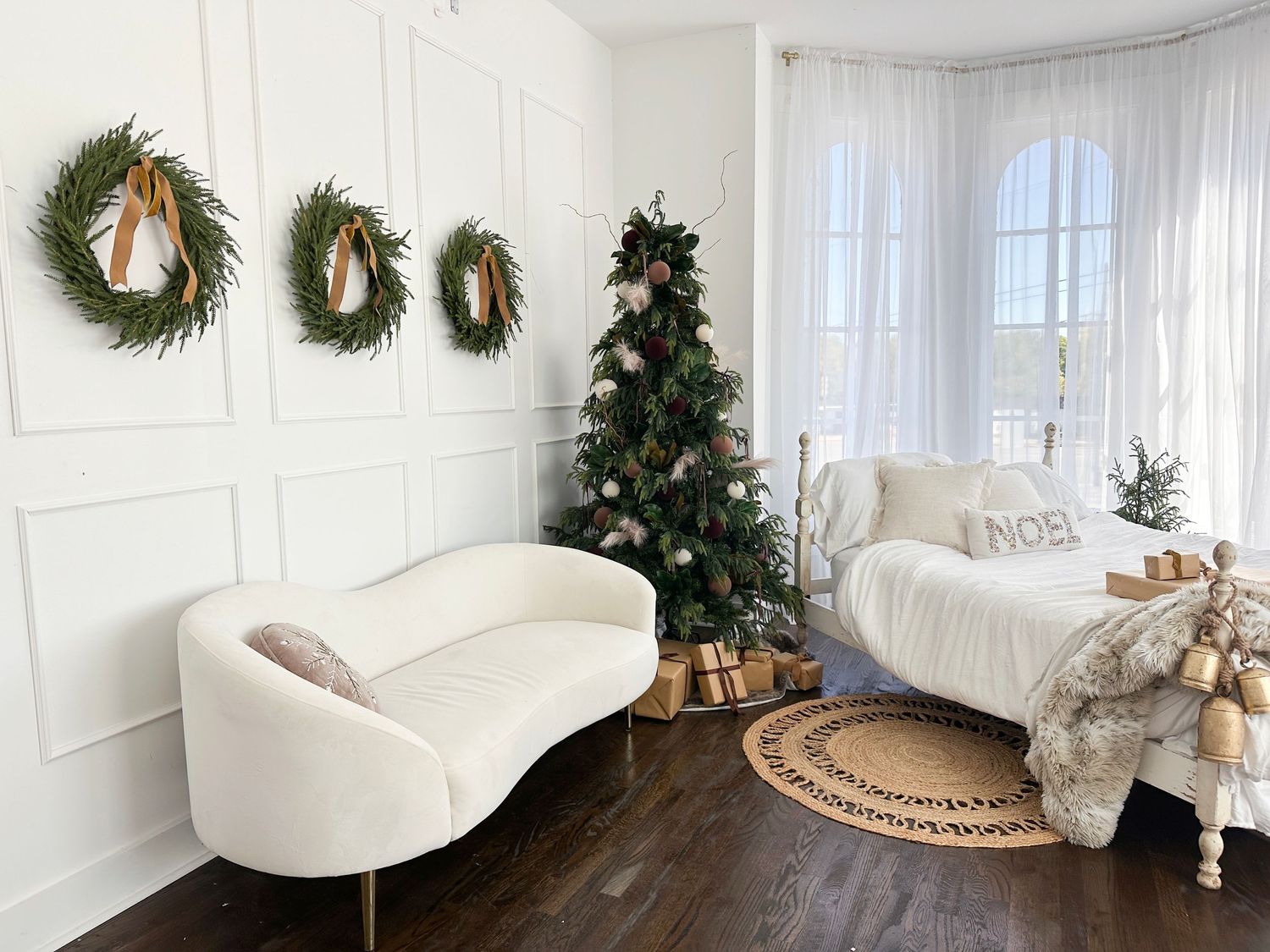 Elegant Christmas living room with white curved sofa, decorated tree, and three wreaths on white wainscoting walls.