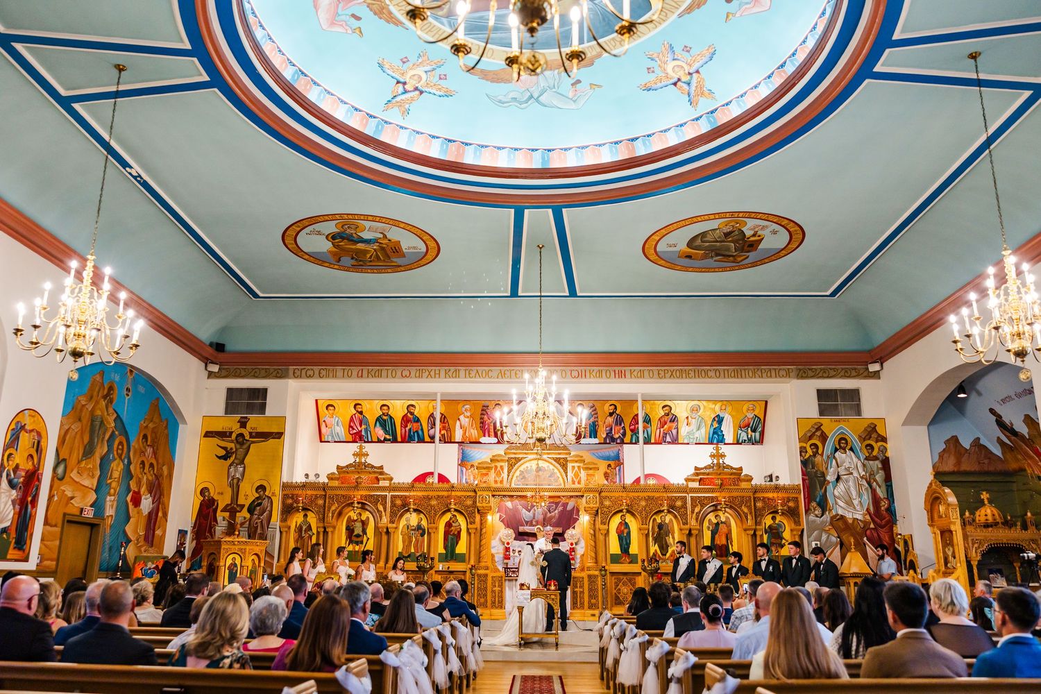 Orthodox church wedding ceremony with ornate golden iconostasis, colorful domed ceiling, and guests seated in pews.