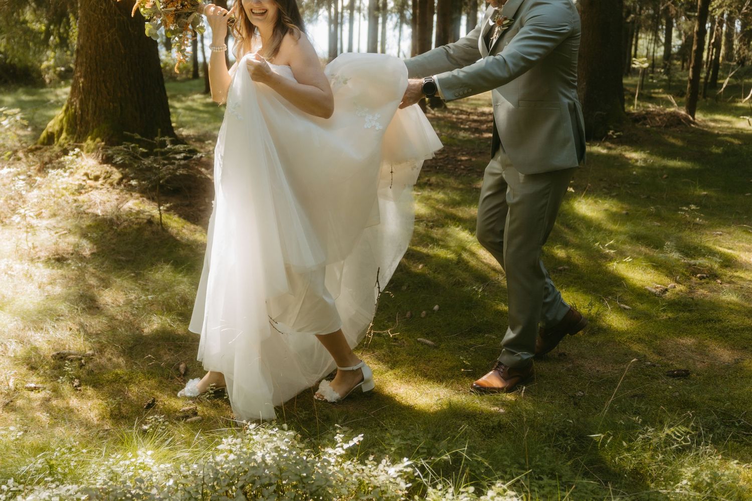 Bride and groom dancing outdoors in sunlit forest, white wedding gown flowing in breeze, groom in gray suit.