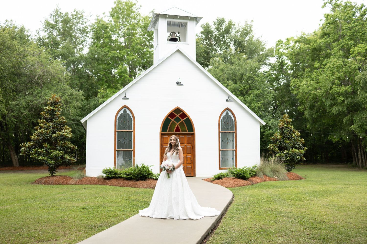 Bride in white gown poses before a charming white chapel with arched windows and wooden doors on a lush green lawn.
