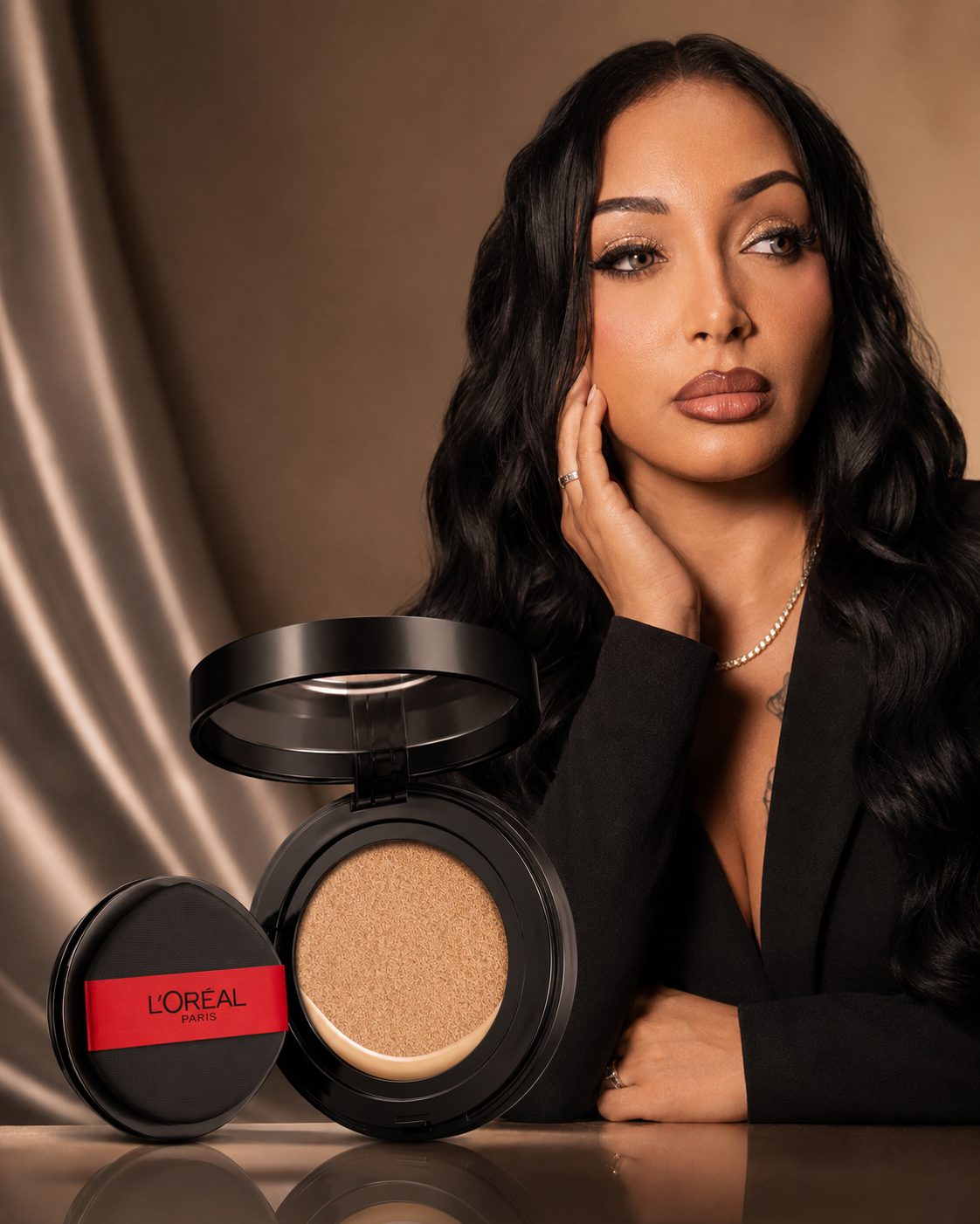 Woman with long wavy black hair in a black blazer poses beside an open L'Oréal cushion foundation compact against a warm bronze backdrop.