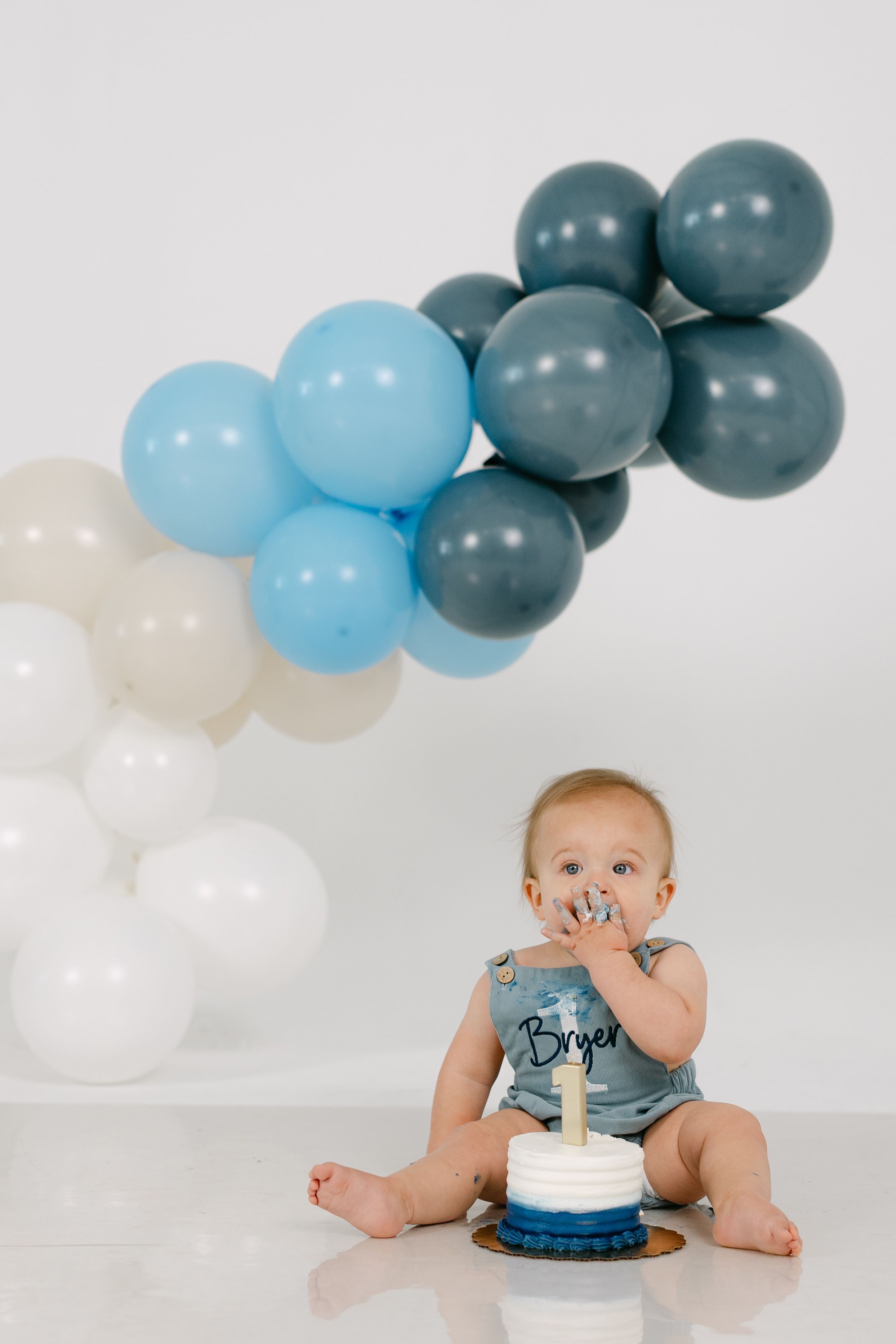 Baby in a blue romper named Bryer eating a smash cake with a blue and white frosted tier beneath a blue, gray and white balloon garland.