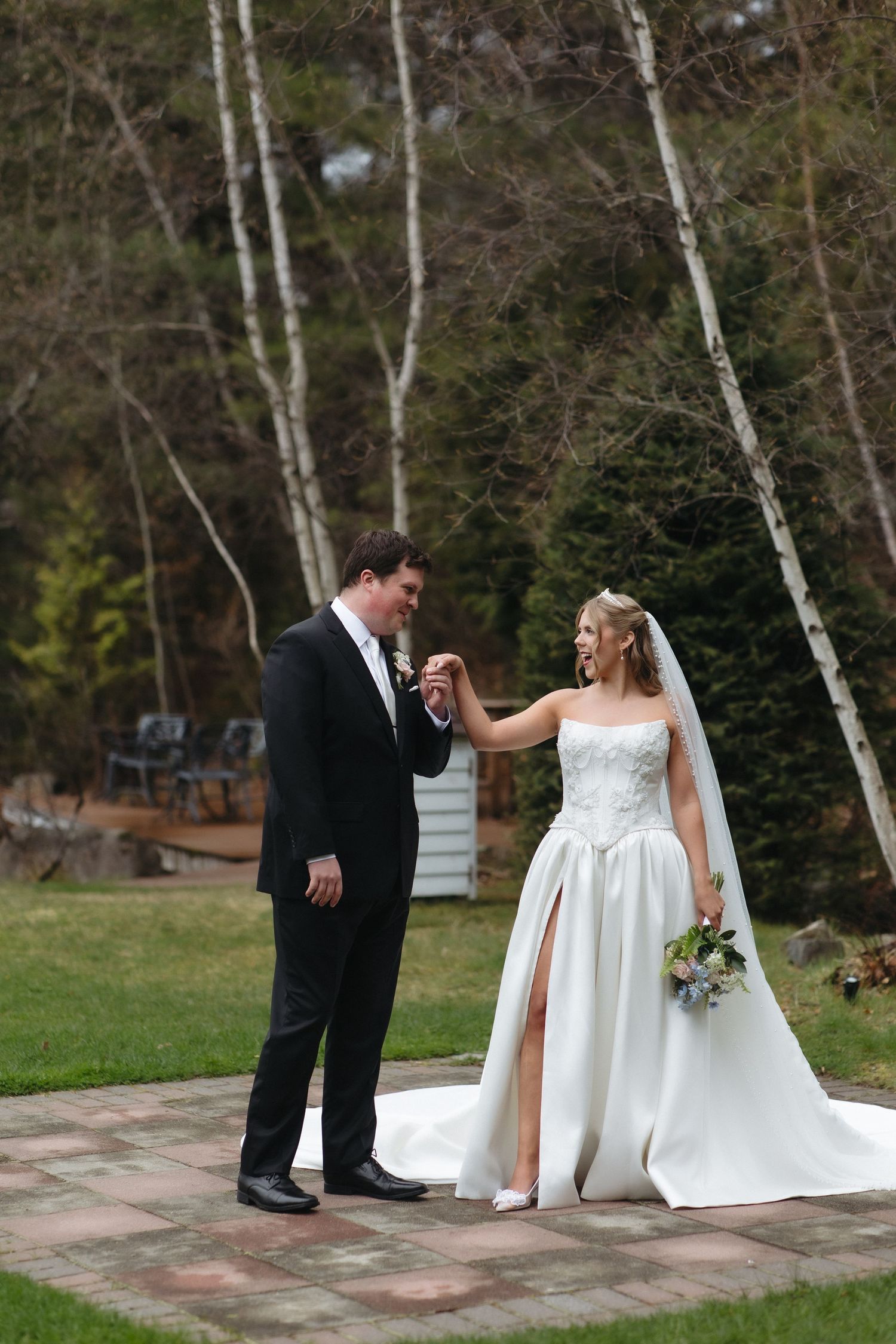 Bride and groom hold hands on brick path outdoors, she in white gown with bouquet, he in black tuxedo, birch trees behind.