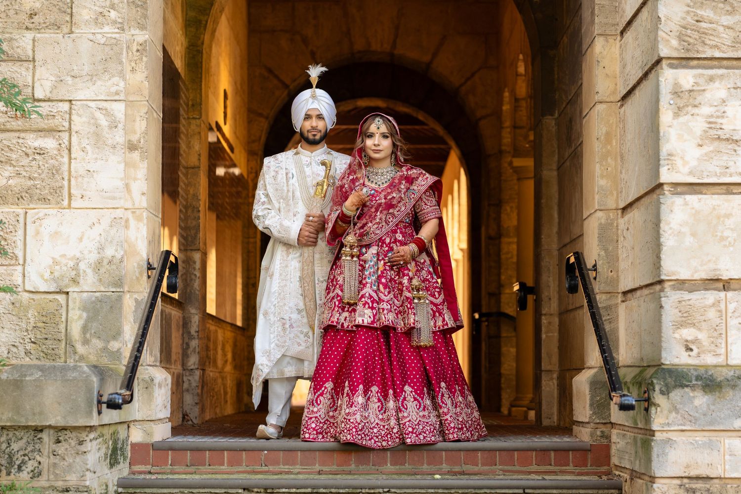Indian bride and groom in traditional red and white wedding attire posing under stone archway.