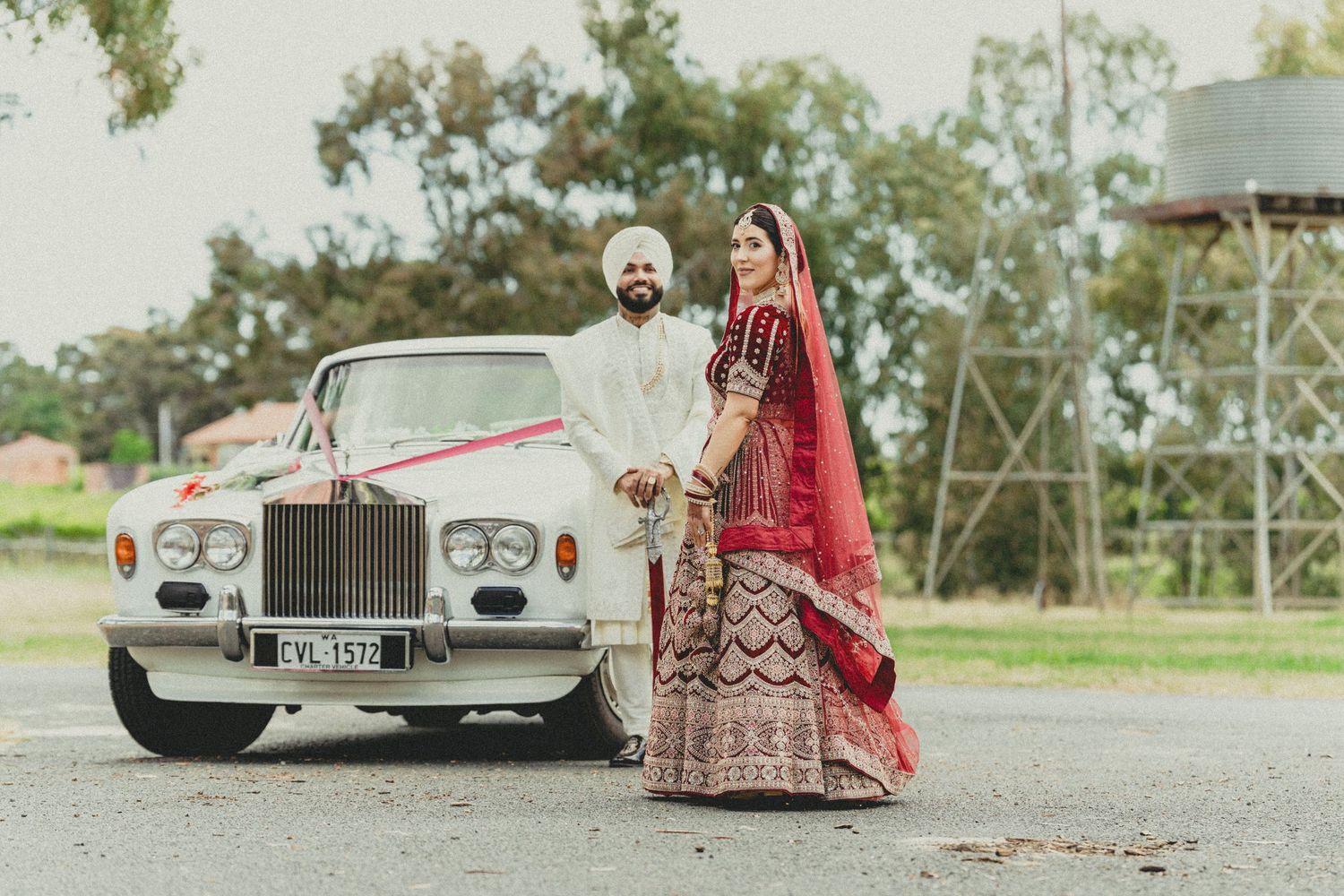 Sikh bride and groom in traditional attire posing beside a classic white Rolls-Royce decorated with red ribbon.