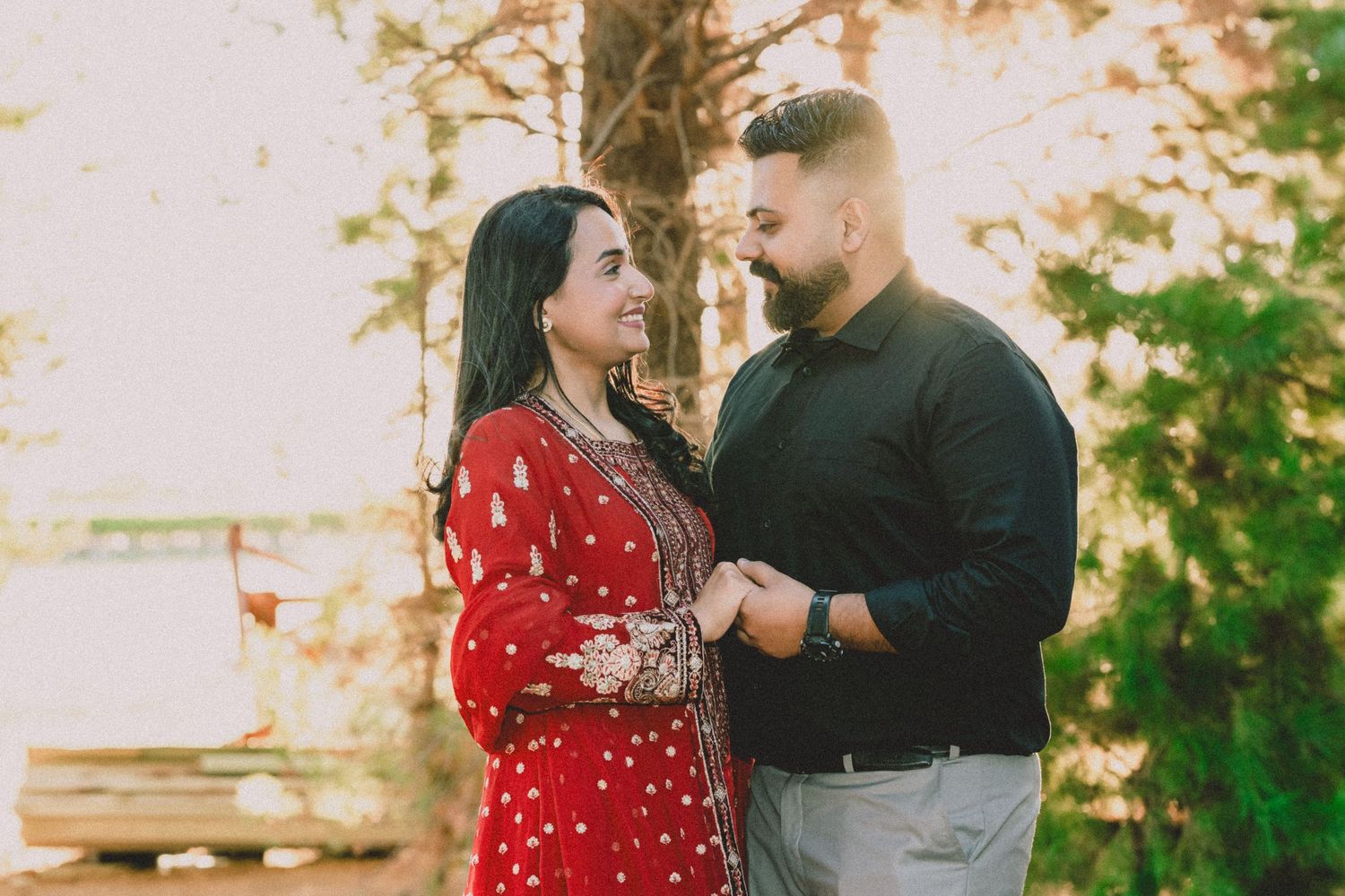 Couple in traditional South Asian attire sharing a romantic moment outdoors, woman in red embroidered dress, man in black shirt.