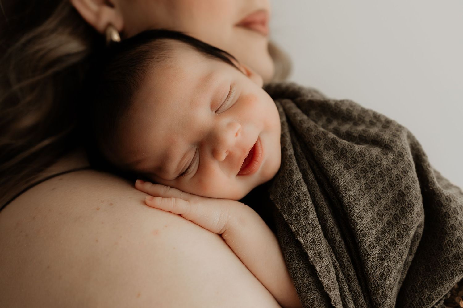 Newborn baby sleeping peacefully on mother's shoulder, wrapped in olive green knit blanket.