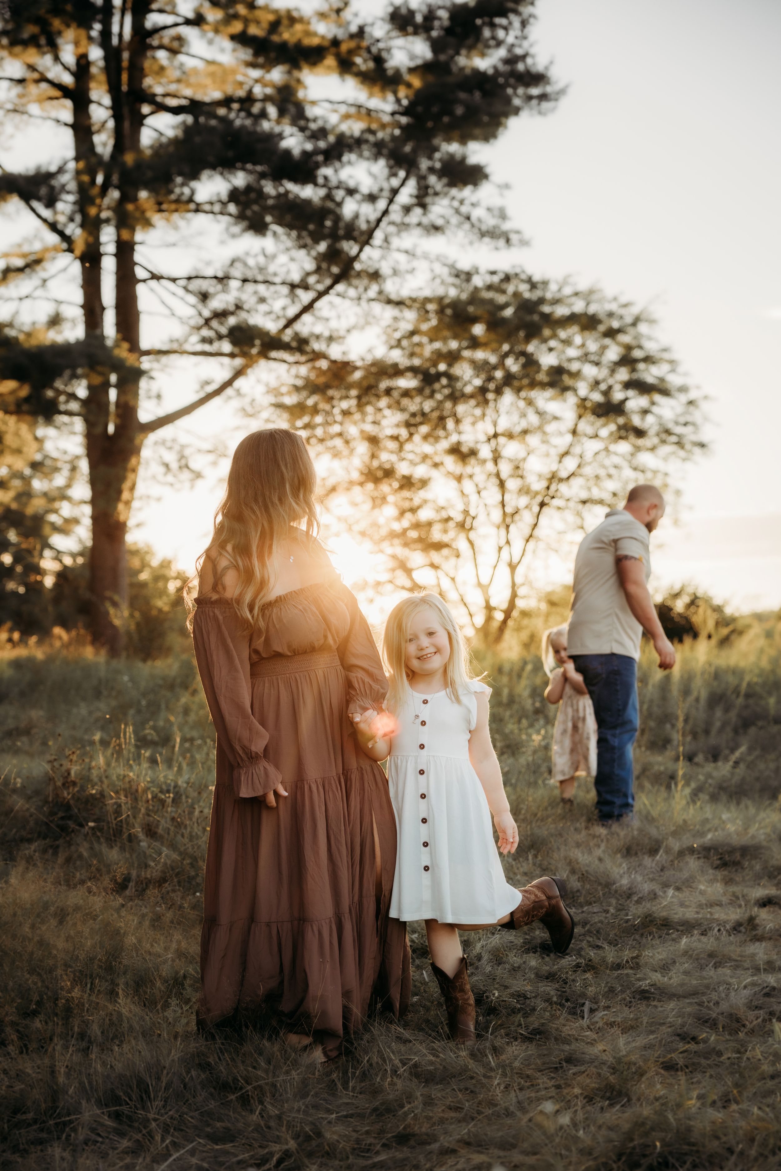 Family portrait at golden hour in a field, mother in brown dress with young girl in white dress, father walking in background.