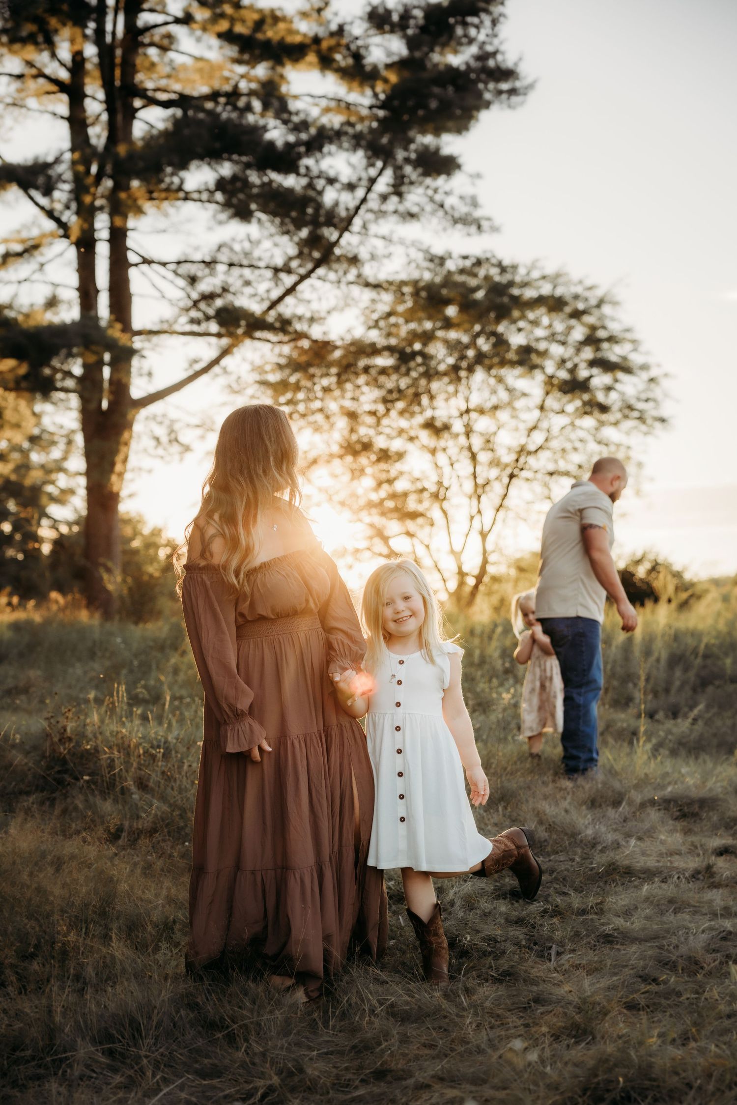Family portrait at golden hour in a field, mother in brown dress with young girl in white dress, father walking in background.