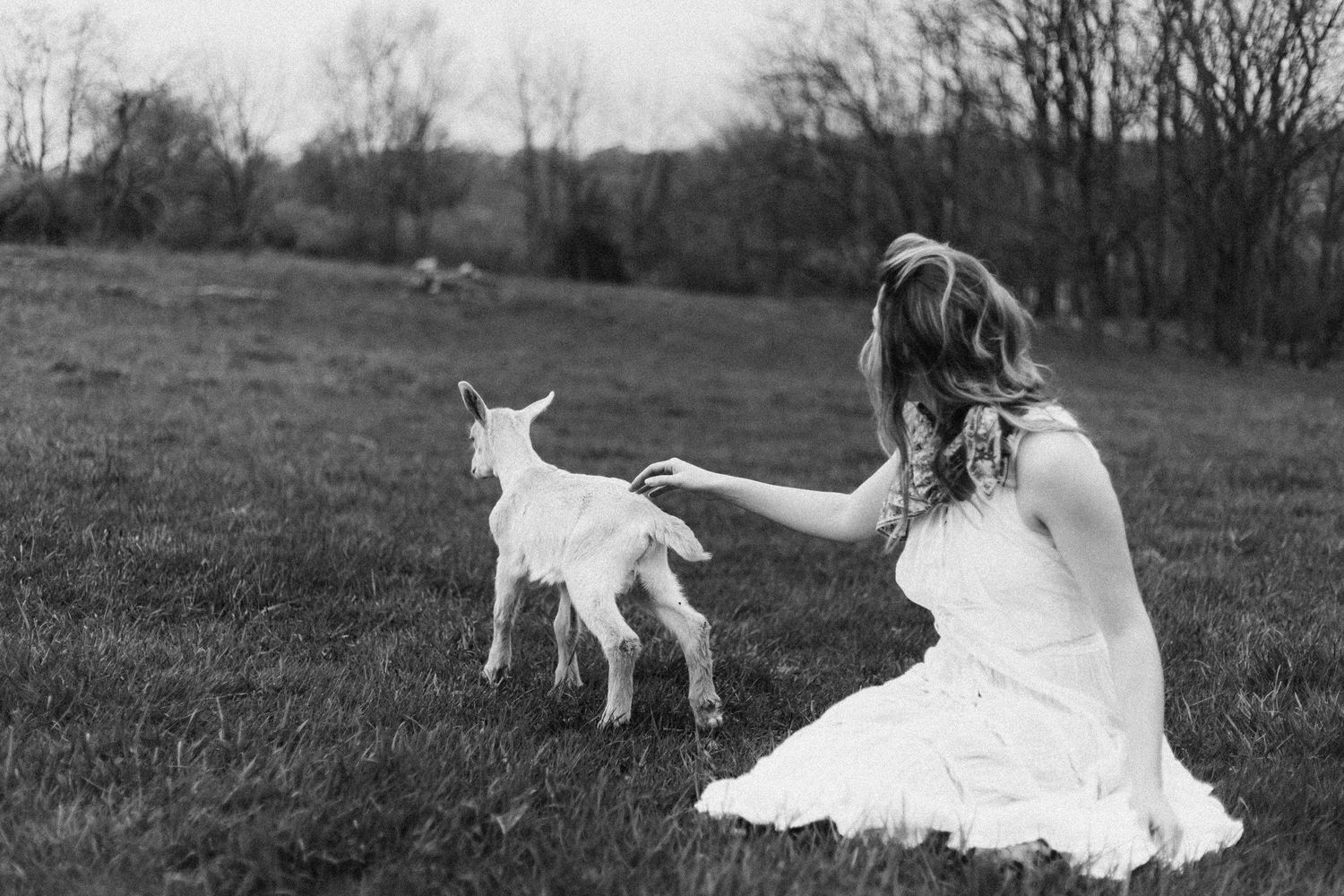Black and white photo of a woman in a white dress kneeling in a field, gently reaching out to a baby goat.