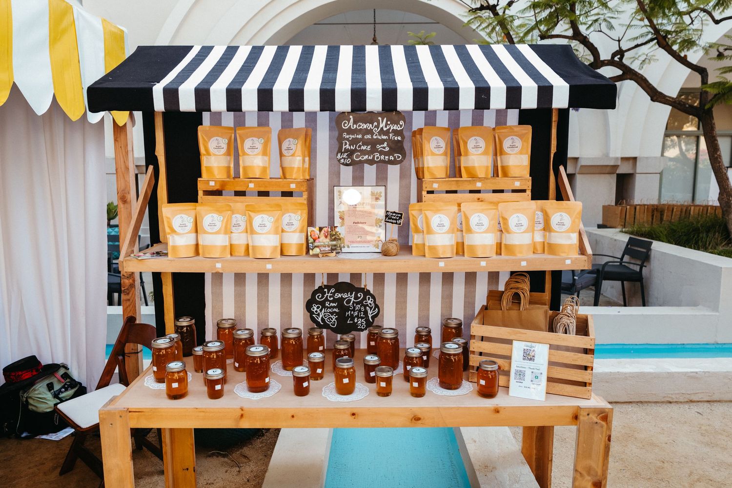 Outdoor honey market stall with jars and packaged products displayed on wooden shelves under striped awning.