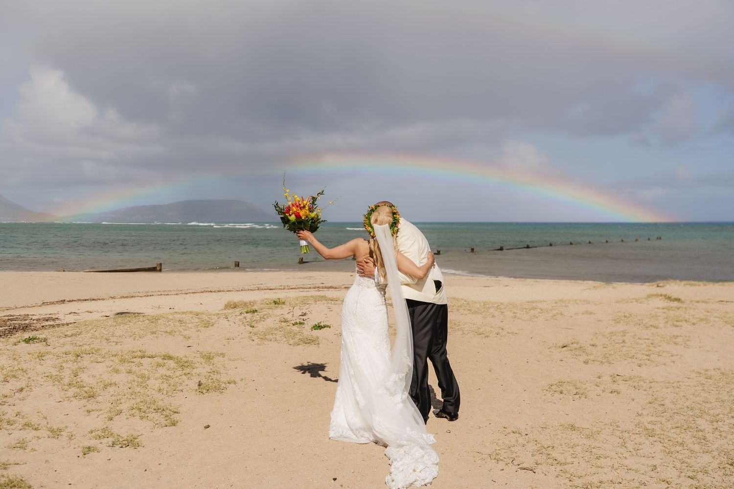Bride and groom kissing on a sandy beach with a vibrant rainbow over the ocean on their wedding day.