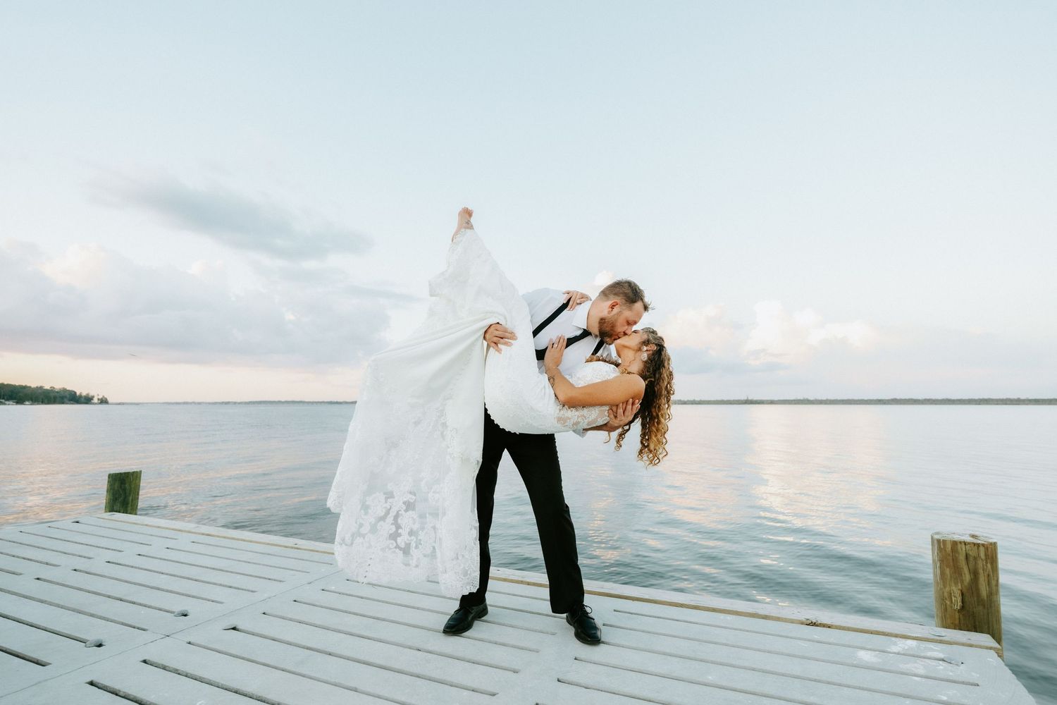 Bride and groom share a romantic dip kiss on a white dock overlooking calm water at sunset.