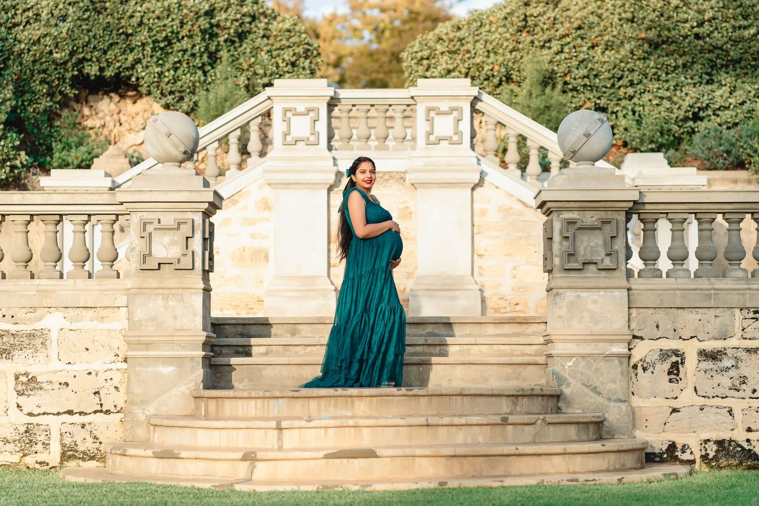 Woman in a green dress sits peacefully in an outdoor garden surrounded by white floral arrangements in gold urns on a brick patio.