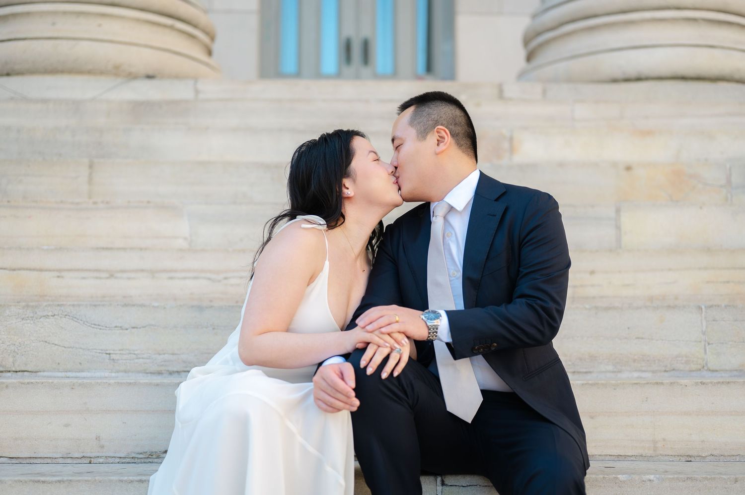Newlywed couple in a white wedding dress and navy suit sharing a kiss while sitting on stone steps after their elopement in Brooklyn.