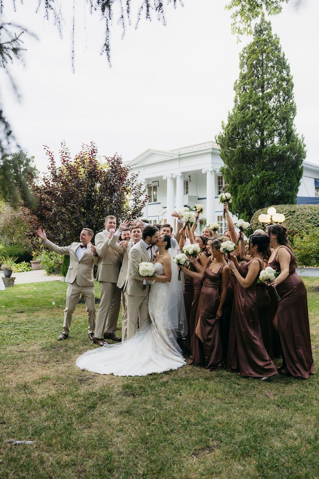 Bride and groom embrace before a grand white colonial mansion with tall pillars and colorful flowers at their wedding.