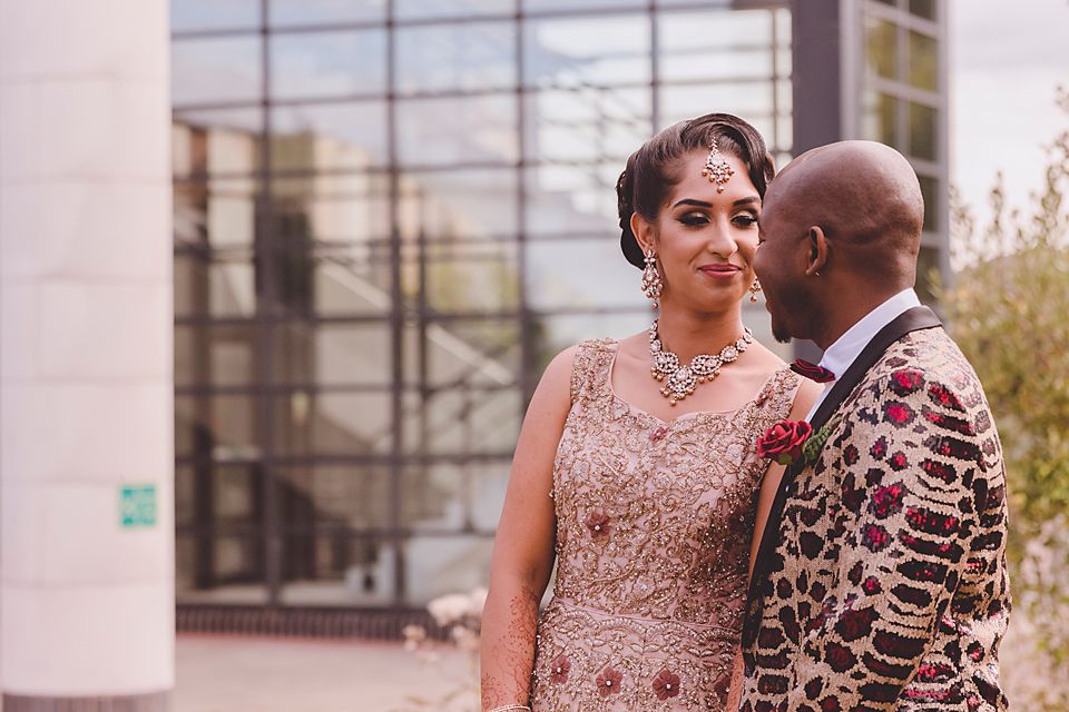 Couple in ornate formal attire embracing among tall pampas grass at sunset.
