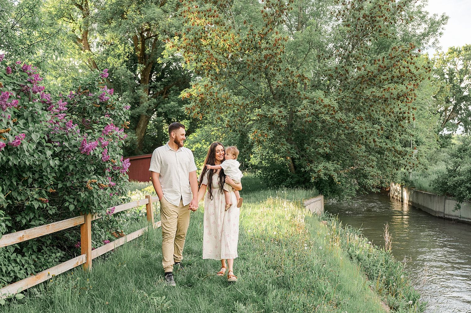 A couple strolls along a grassy path next to a scenic creek with lush greenery in the background.