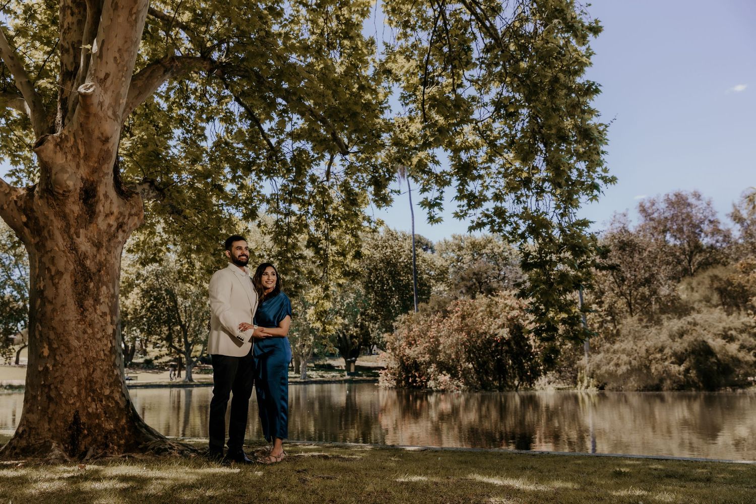 Romantic couple stands under large tree by serene lake during outdoor engagement photoshoot.