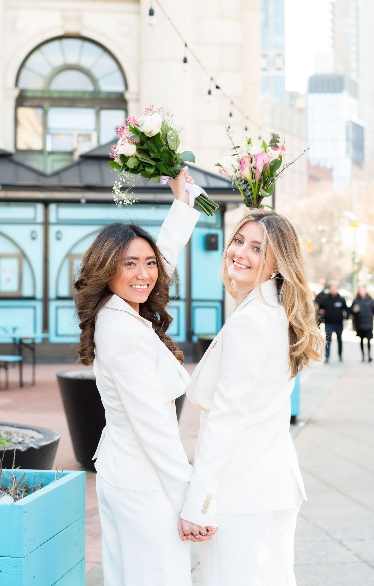 Two people in white outfits pose playfully with flower bouquets against an urban backdrop.