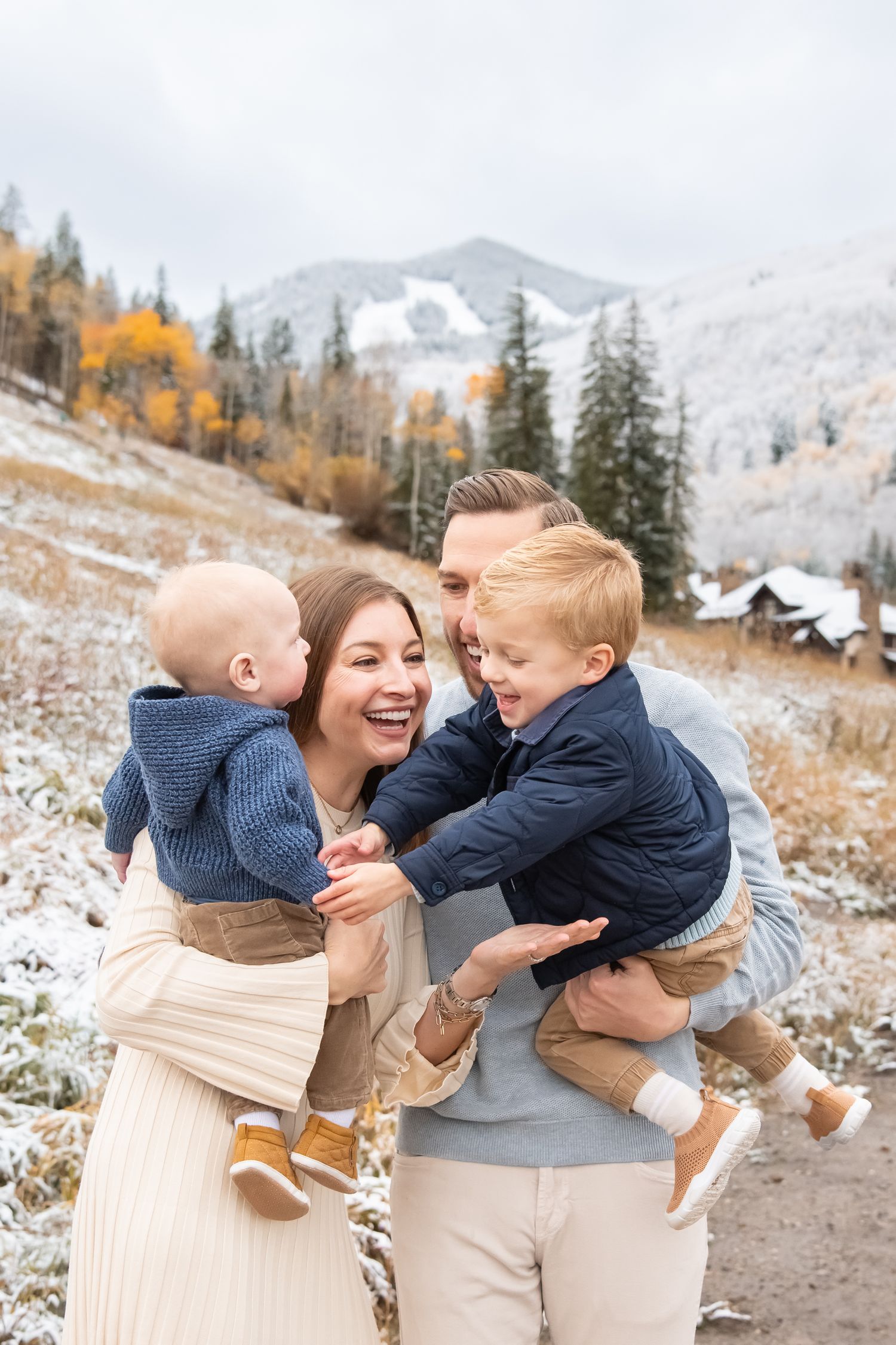 Family portrait session in snowy mountain landscape with golden autumn trees and evergreens in background.