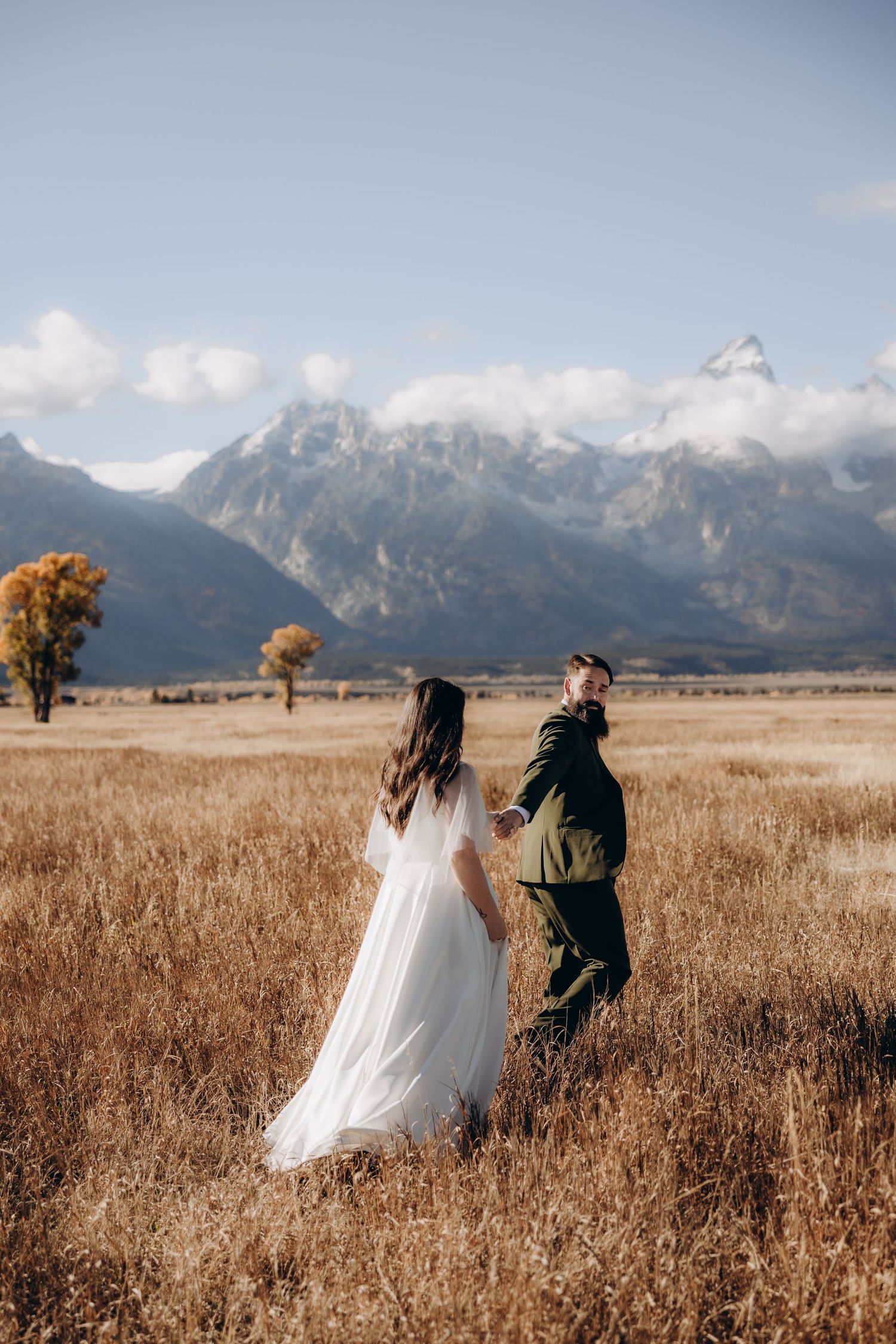 A couple runs playfully through a golden wheat field with majestic mountain peaks in the background.