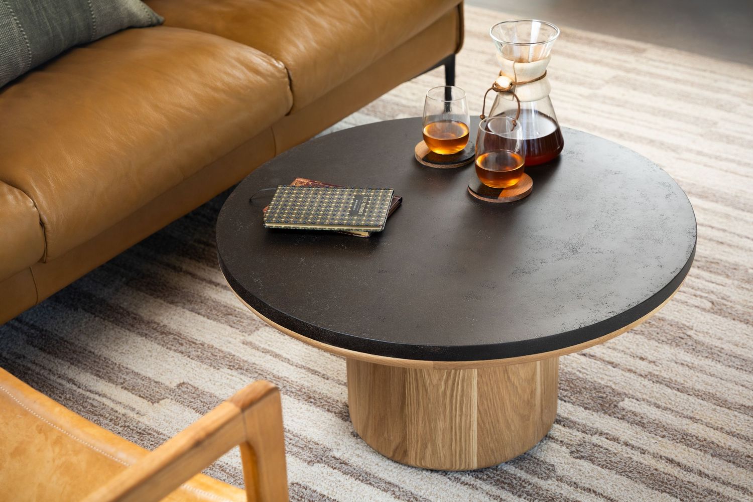 Modern black round coffee table positioned next to a tan sectional sofa on a neutral patterned area rug.