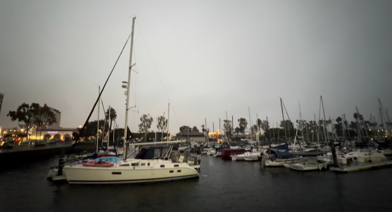 A panoramic view of sailboats anchored in a misty harbor with cloudy skies and distant trees along the shoreline.