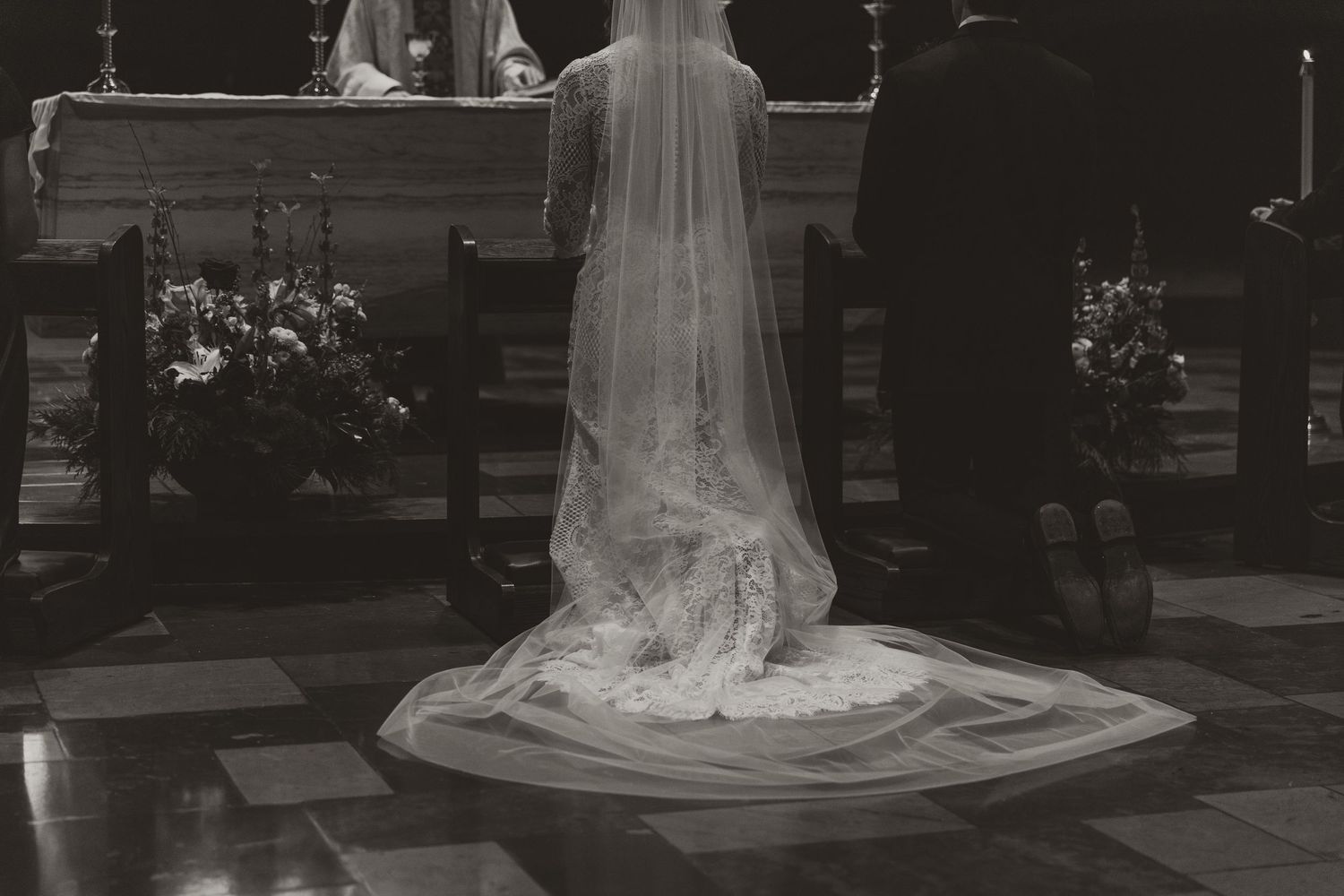 A bride in a flowing white wedding dress and veil walks through a garden pathway in black and white photography.