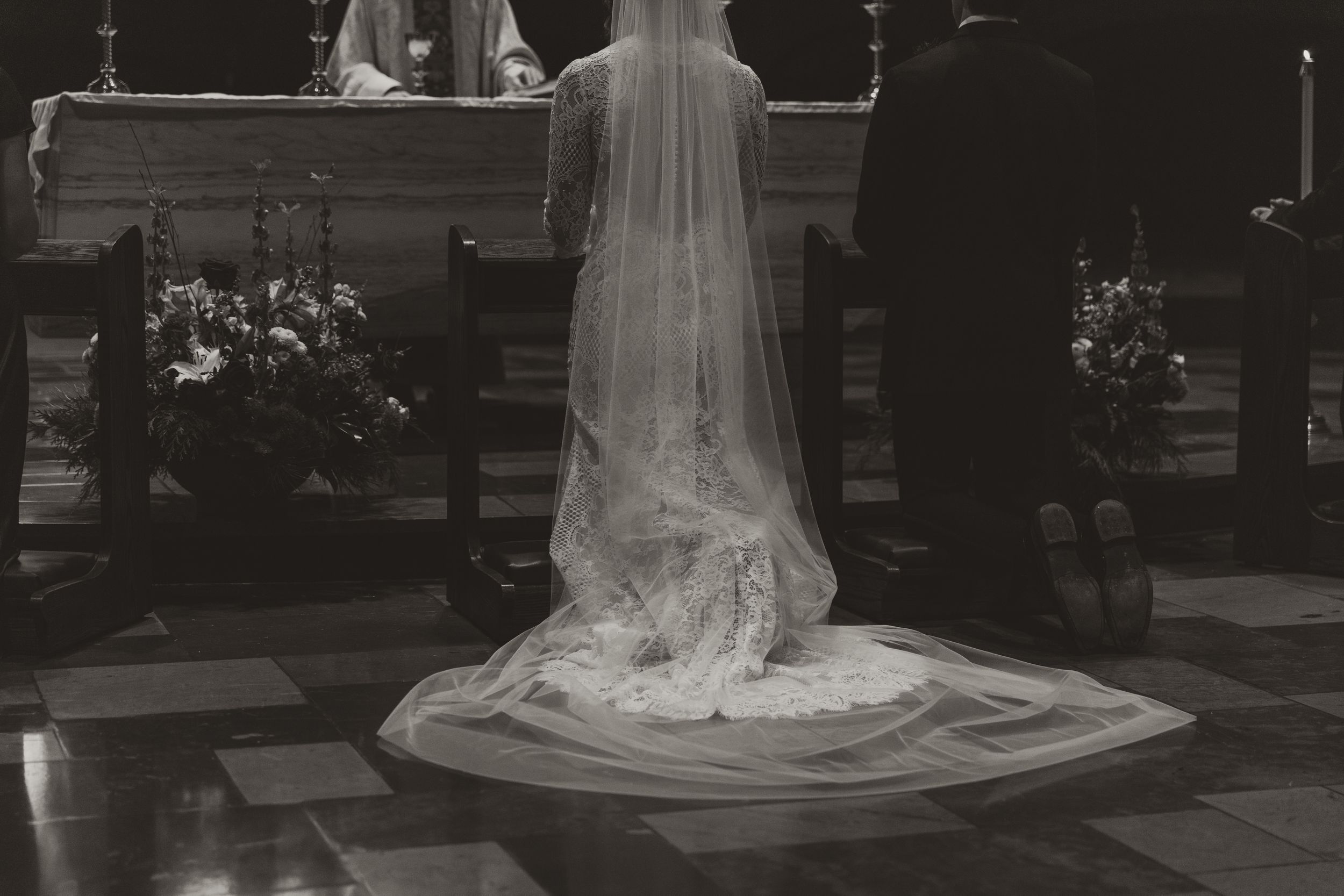 A bride in a flowing white wedding dress and veil walks through a garden pathway in black and white photography.