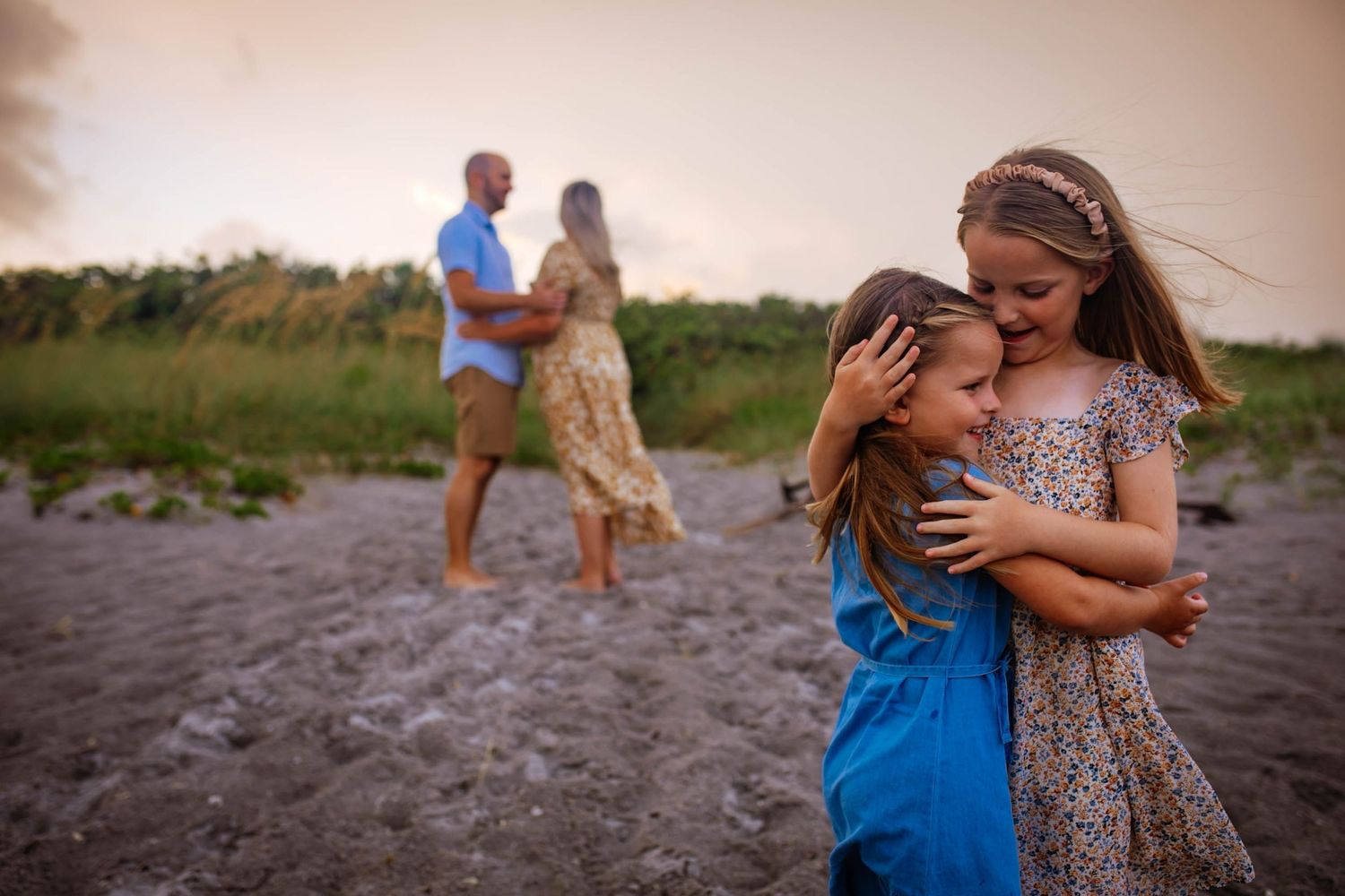 Two children play in the foreground while two people embrace in the distance on a beach during sunset.