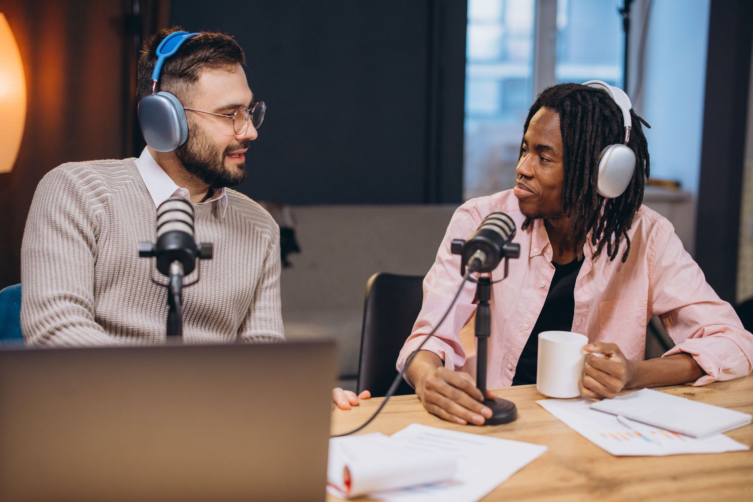 Two people sit at a desk with microphones and headphones while recording a podcast in a studio setting.