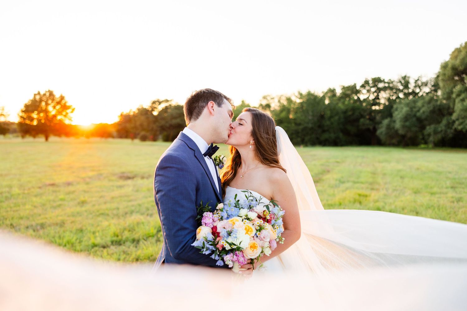 A newlywed couple sharing a romantic moment in a sun-drenched field at golden hour with colorful wedding bouquet.