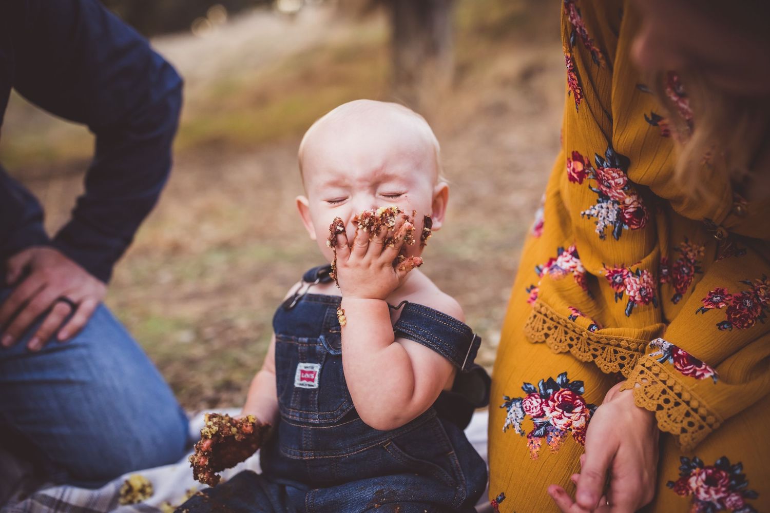 Young child in denim overalls covers face with muddy or chocolate-covered hands while sitting outdoors.