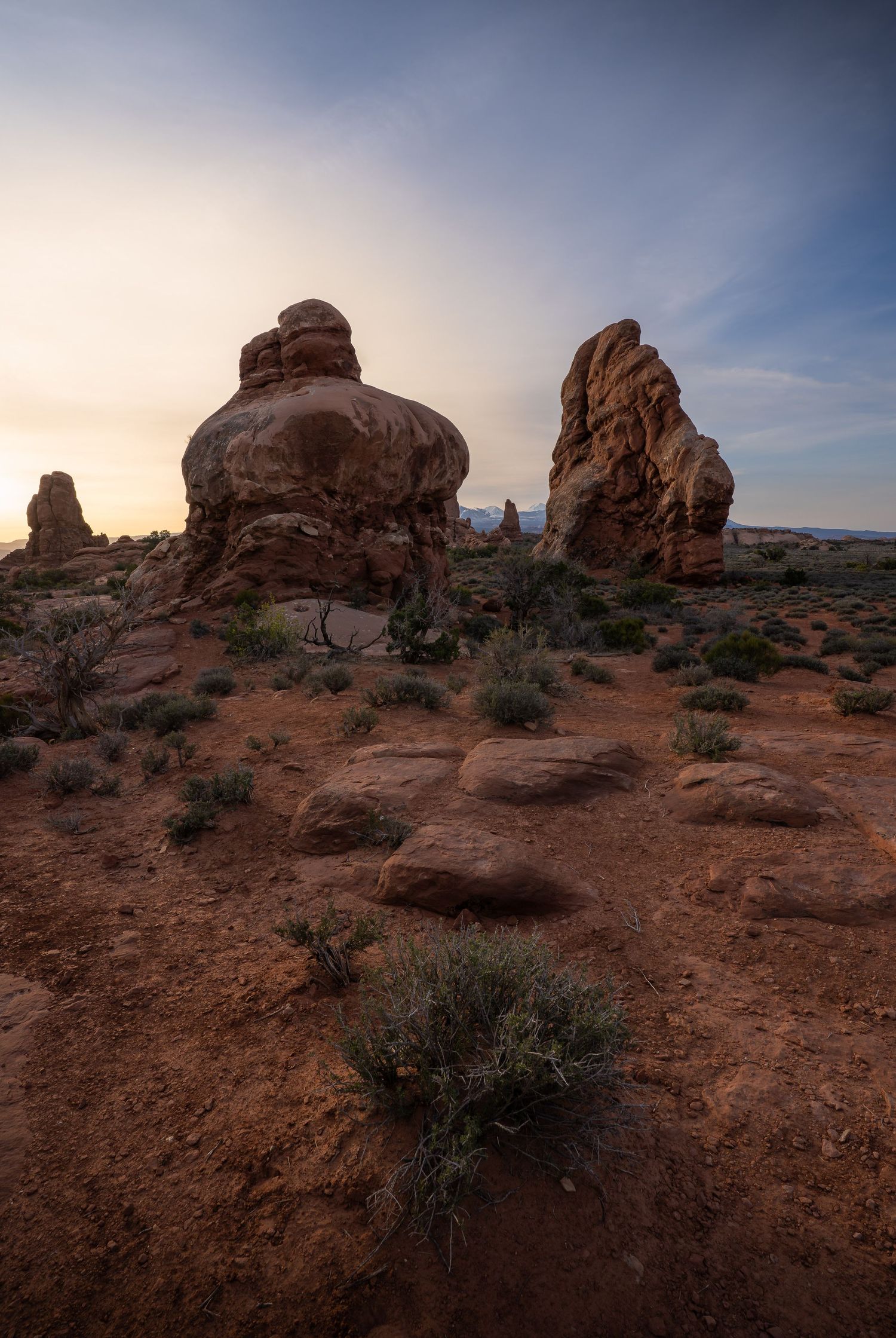 A series of silhouetted figures stand against a sunset sky viewed through natural rock arches in a desert landscape.