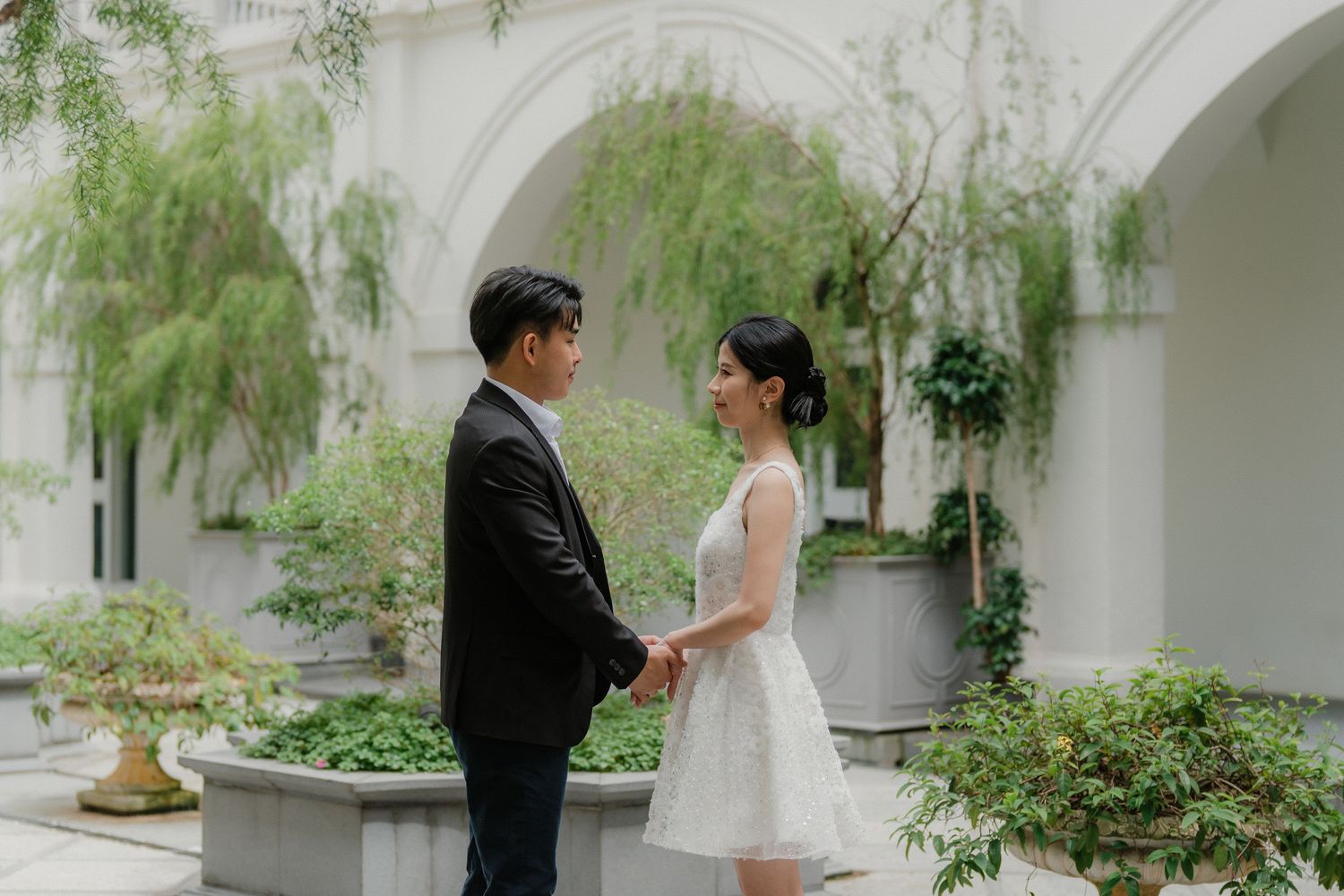A couple stands facing each other in a courtyard garden with white arches and hanging plants in the background.