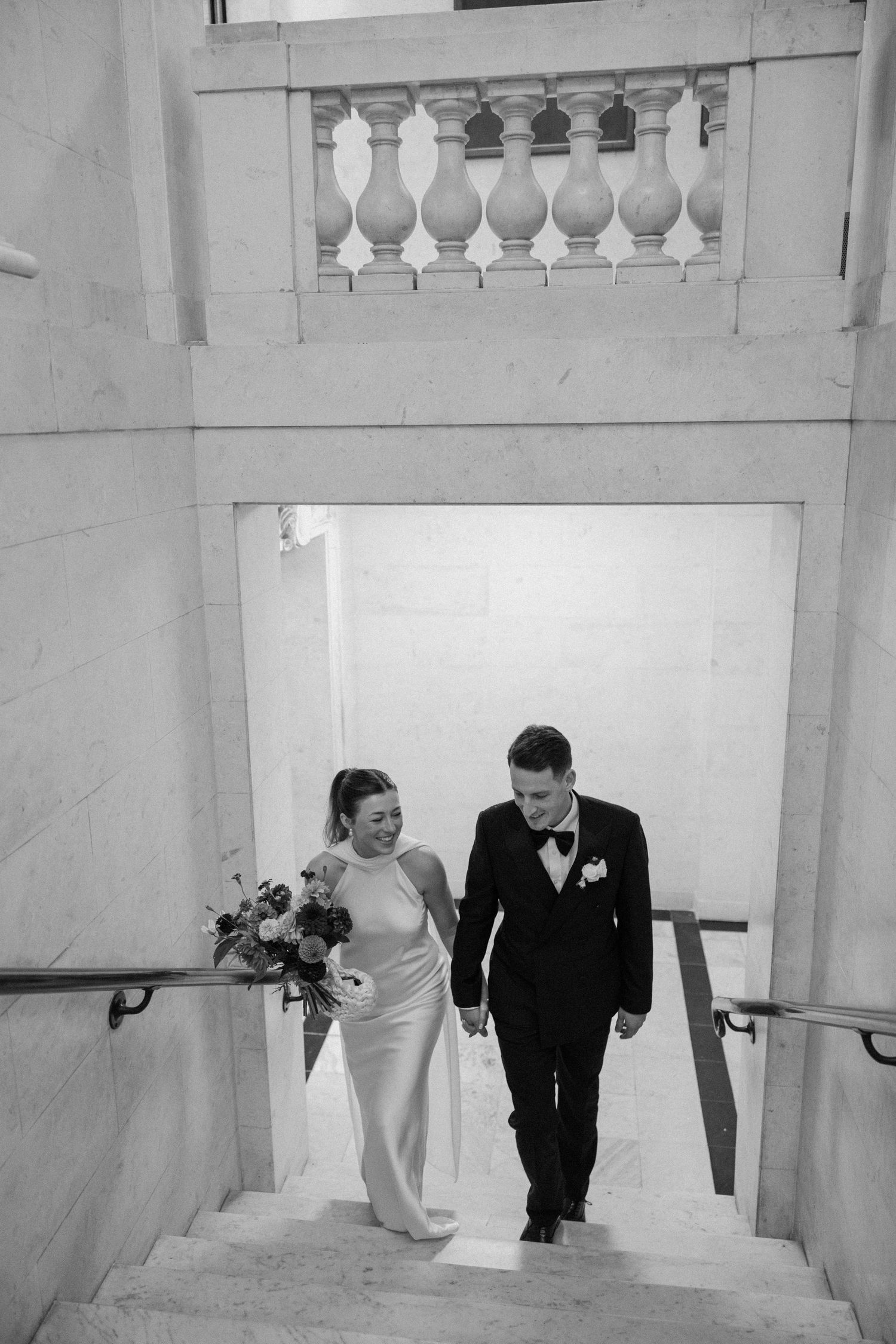 A black and white sequence shows a couple ascending marble stairs in formal wedding attire at Old Marylebone Town Hall in London.