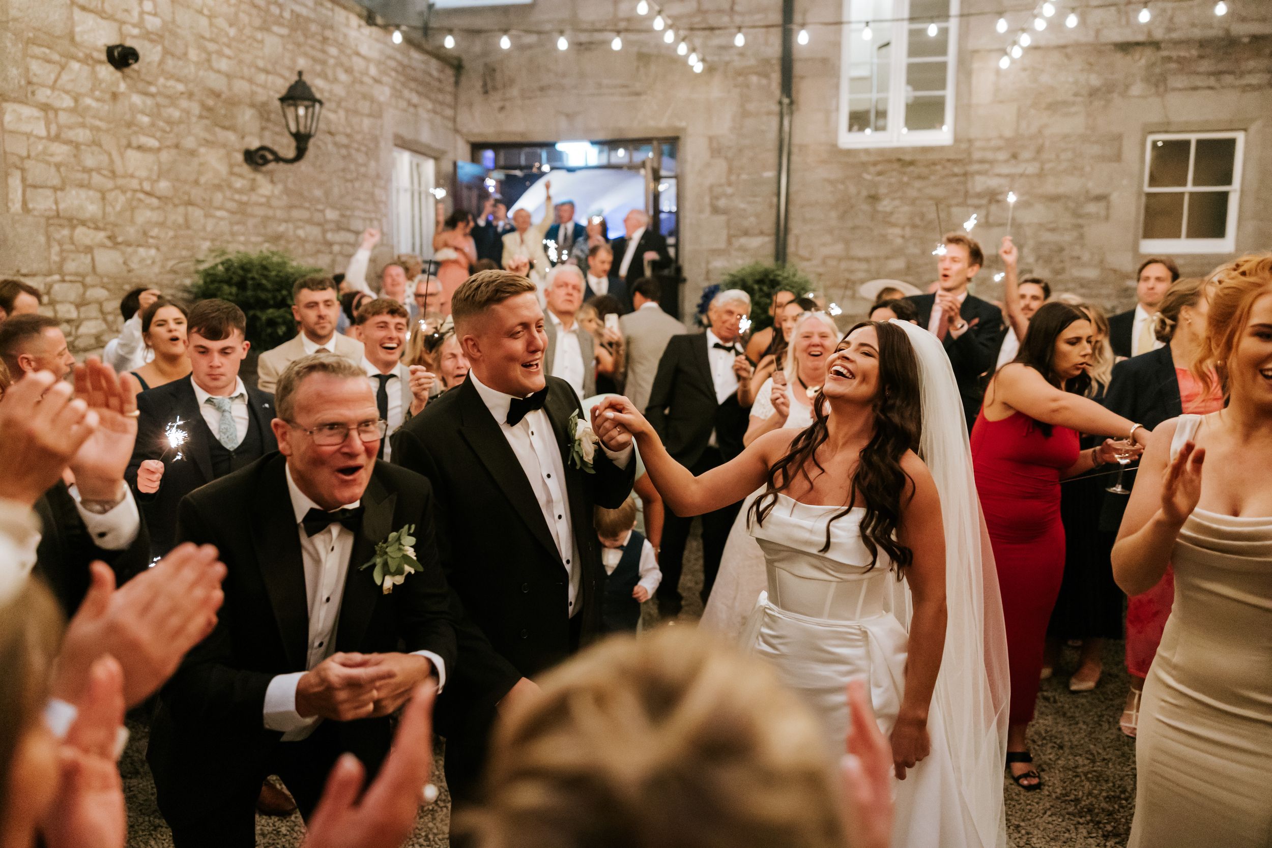 Wedding guests dance and celebrate together under string lights in a rustic stone-walled reception venue.