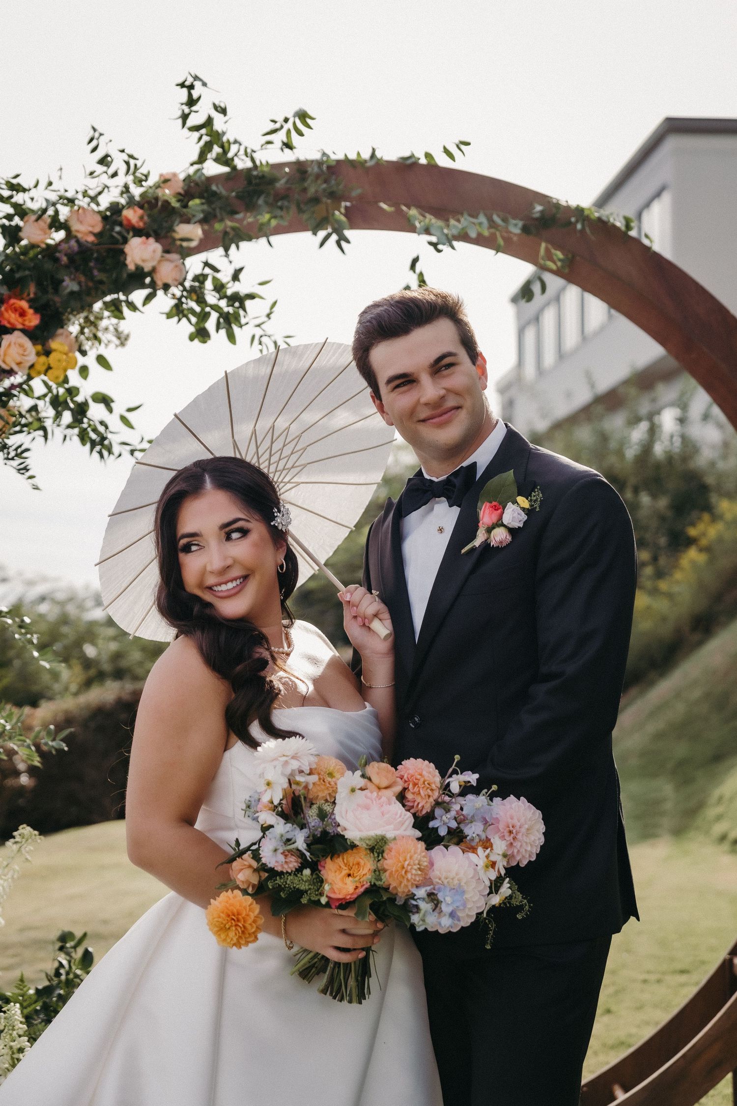 Romantic wedding couple poses outdoors with white parasol and vibrant orange and pink floral bouquet.