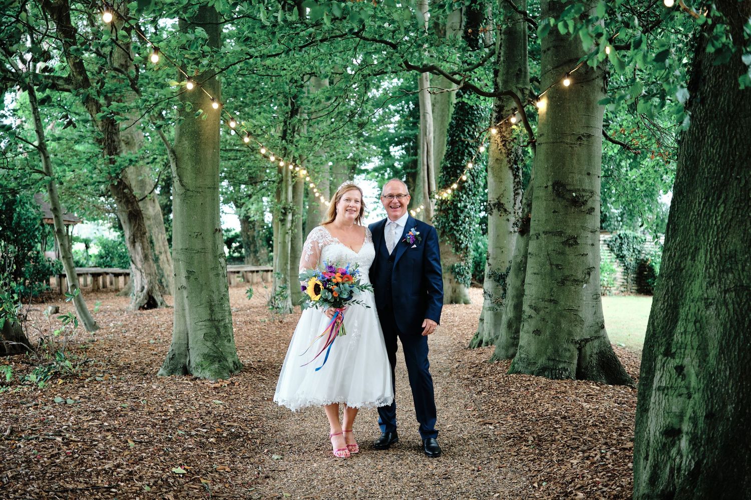 Couple walking through a tranquil wooded area in formal attire on a trail lined with tall trees outside The Crown Lodge in Wye, Kent.