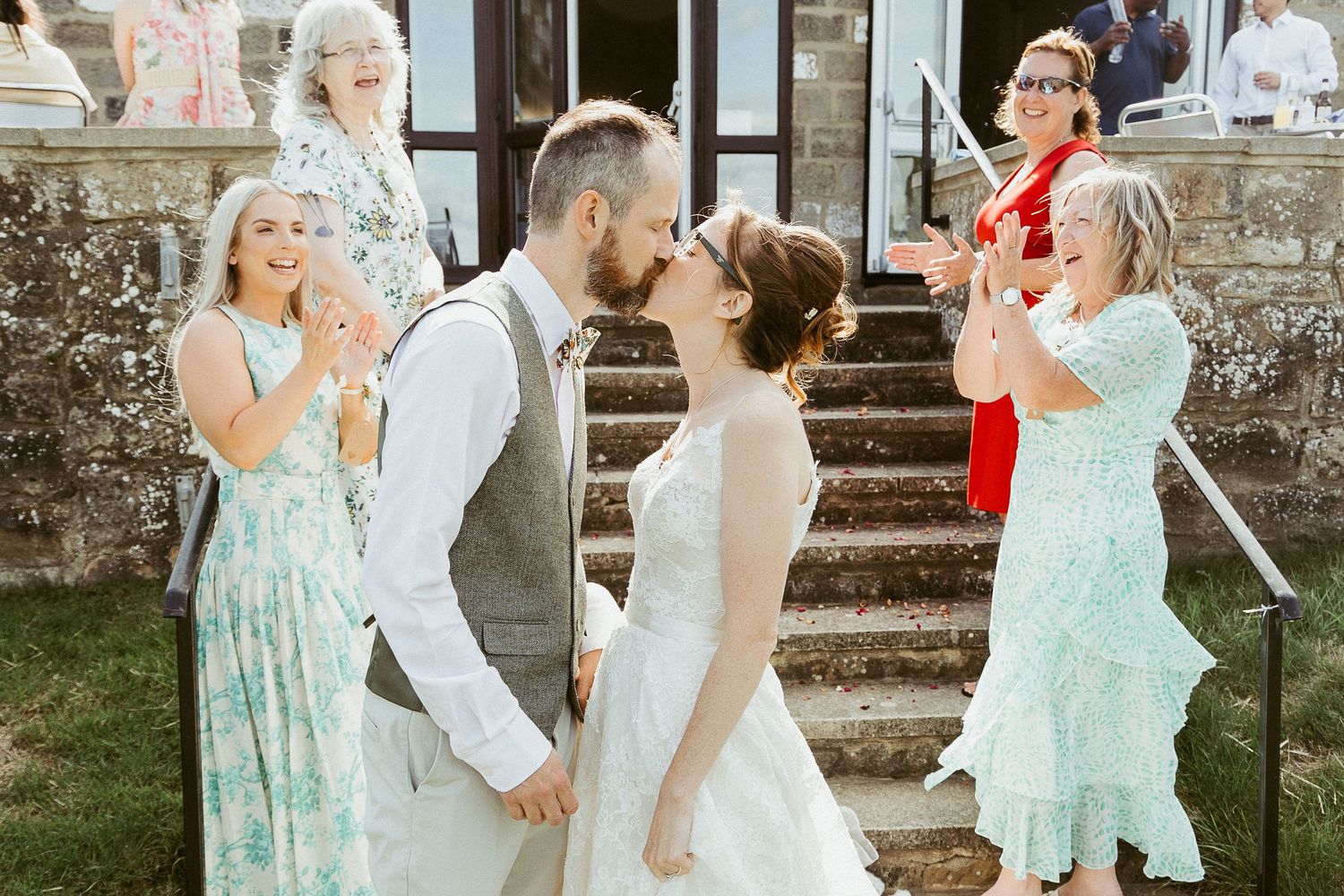 Bride and groom sharing a kiss outside East Keswick Village Hall, surrounded by family and friends cheering during their boho Yorkshire wedd