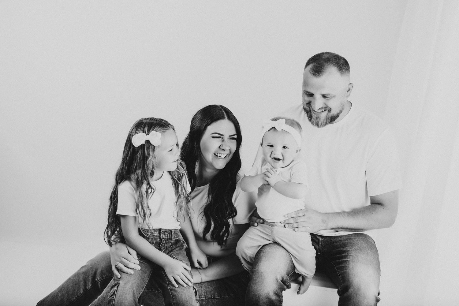 A black and white portrait of a family of four sitting together against a plain background, smiling and laughing.