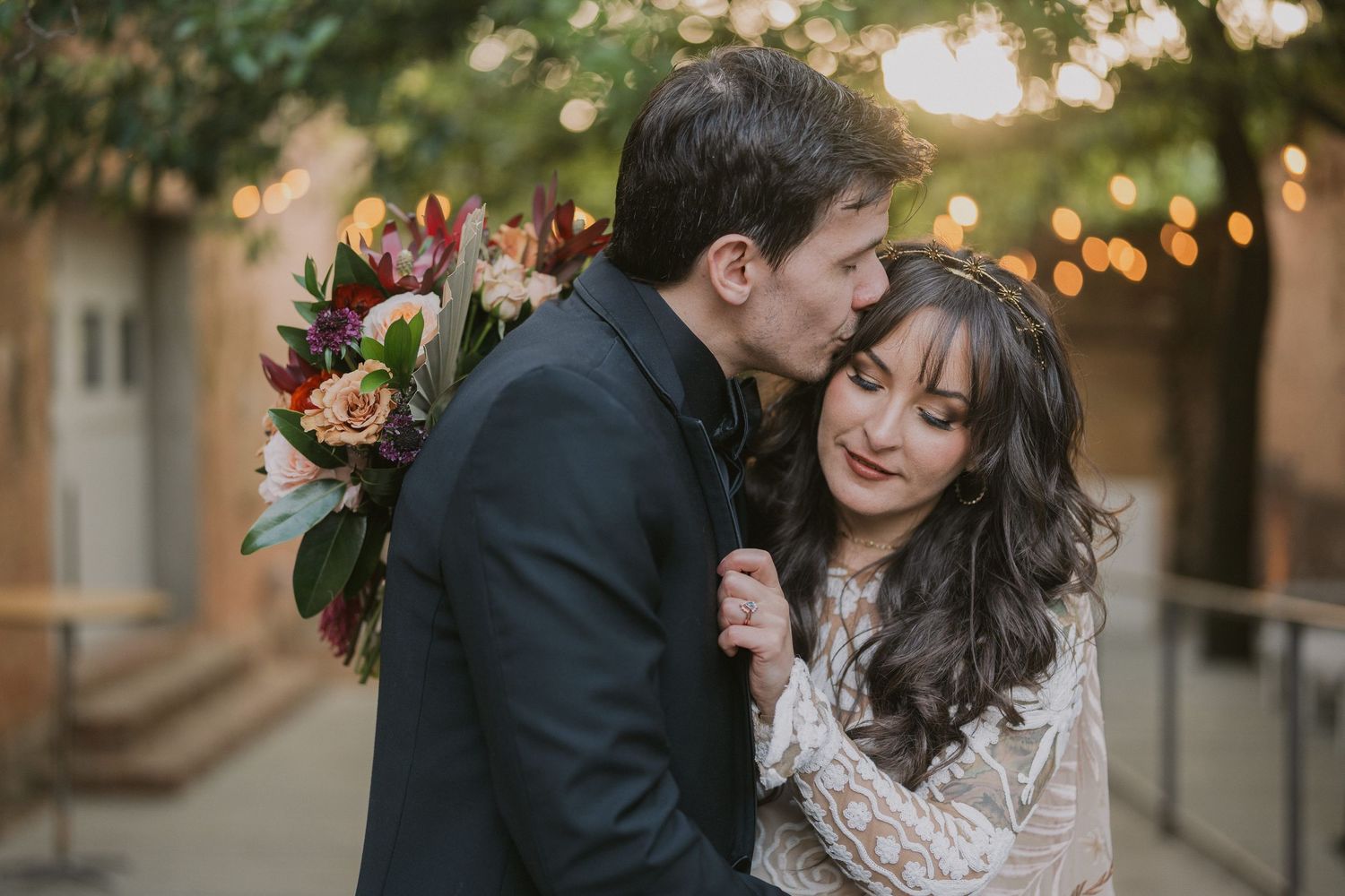 A romantic couple embraces during an outdoor wedding photoshoot with string lights and autumn decor in the background.