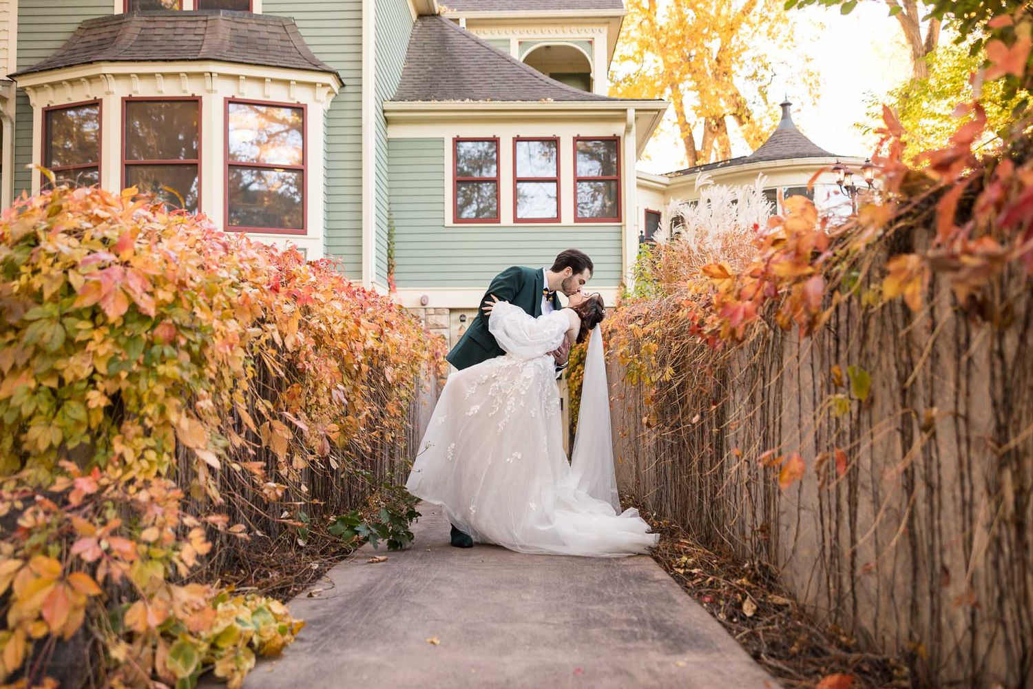 A romantic autumn wedding moment on a walkway with colorful fall foliage and a mint green house in the background.