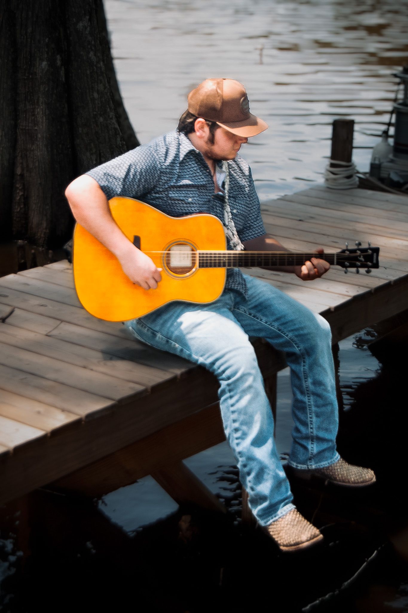 Musician in denim and baseball cap plays acoustic guitar while sitting on wooden dock by water.
