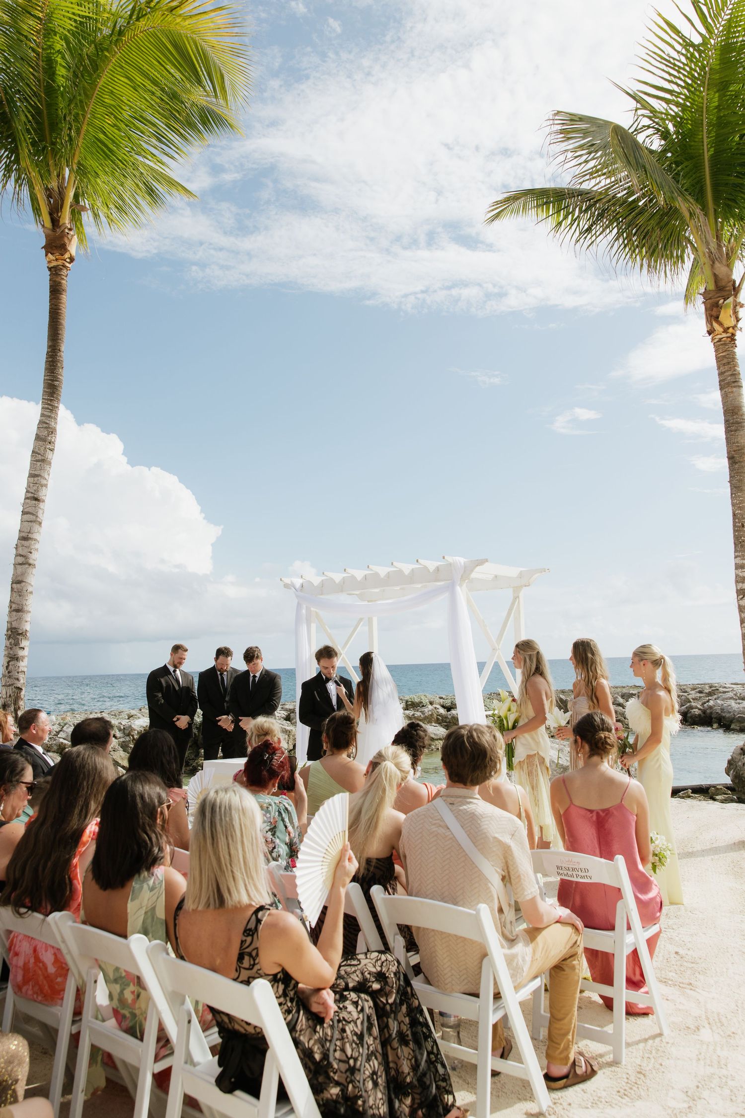 Tropical beach wedding ceremony with guests seated facing ocean view.