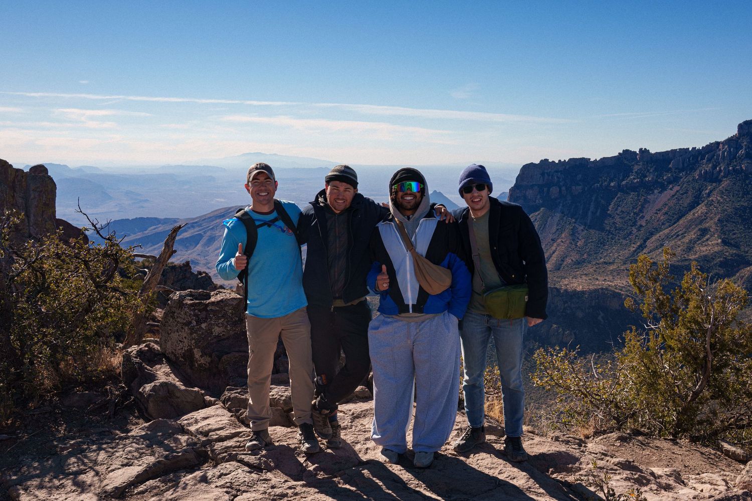 A group of hikers pose together on a mountain summit with scenic valley views and blue skies in the background.