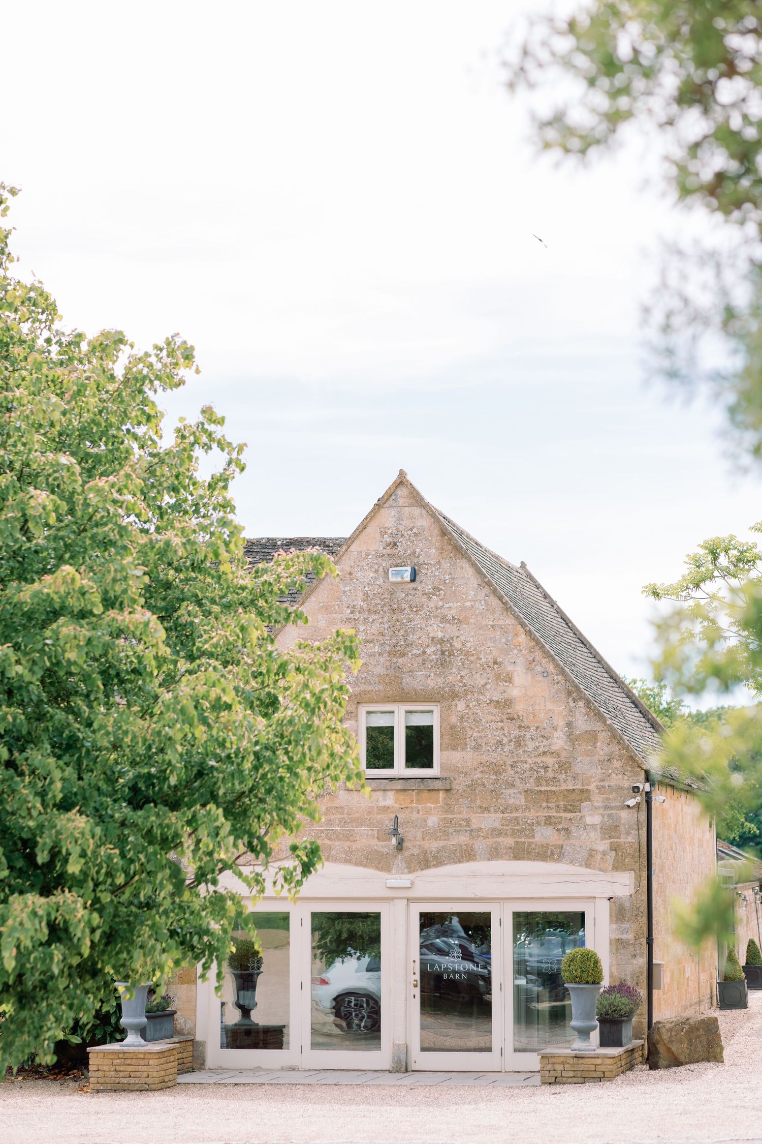 Stone cottage building with white pillared entrance and large window surrounded by lush green trees in soft daylight.