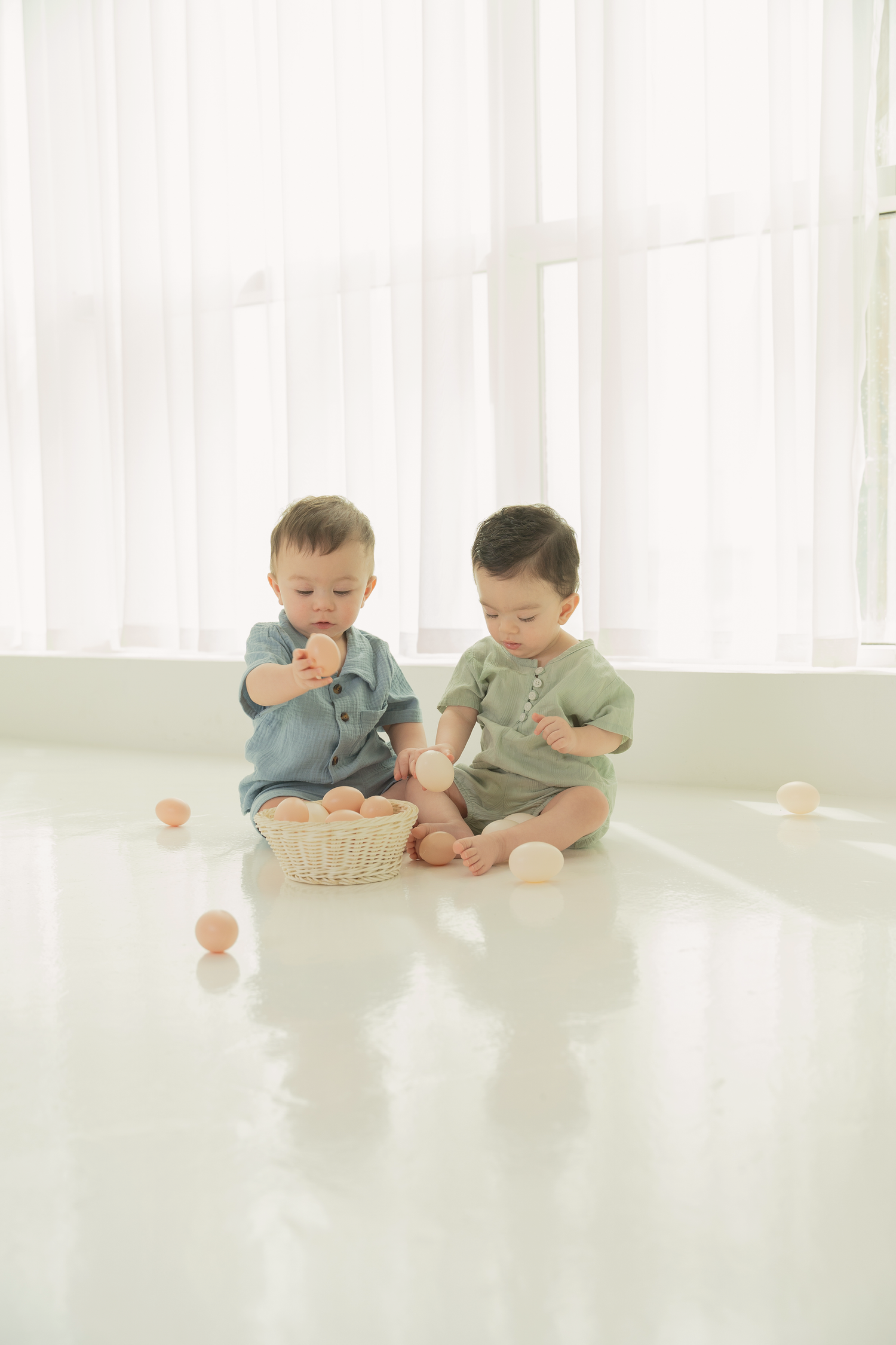 Two babies in matching outfits sit on a white floor with a basket between them and scattered eggs around.